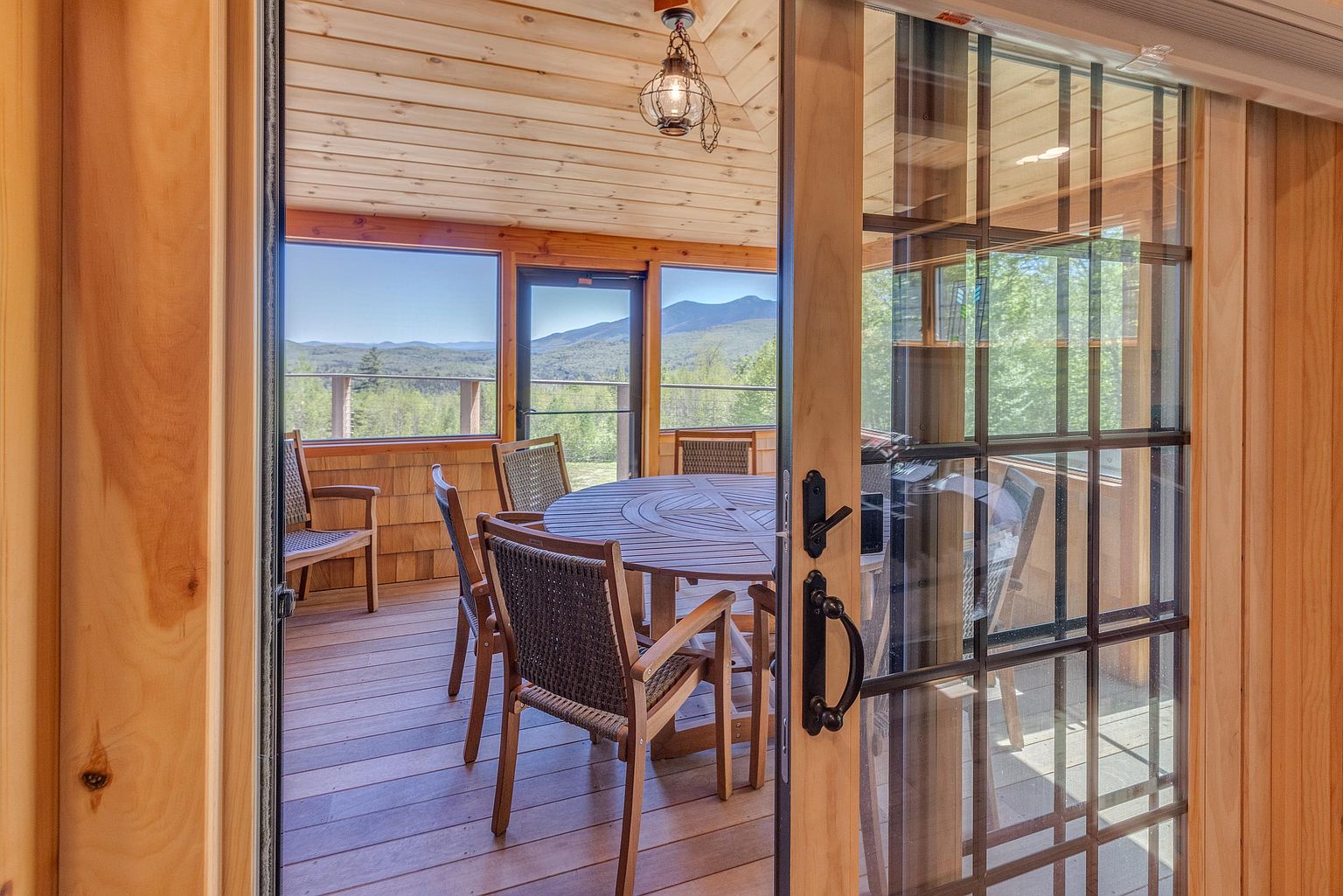 This view captures a screened-in porch from the perspective of an interior room, looking through a glass-paned door. The porch features light wood paneling on the walls and ceiling, a round dining table with chairs, and expansive views of distant mountains. A decorative hanging lantern adds a rustic touch to the space, creating an inviting atmosphere for outdoor dining.