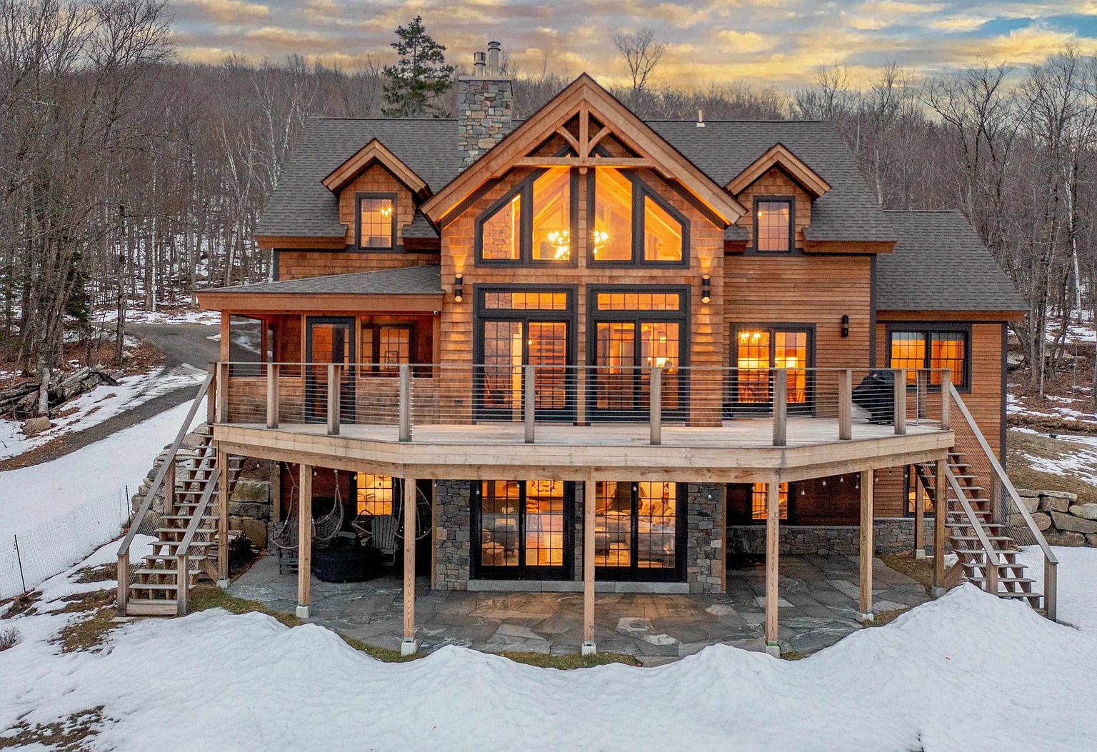 This stunning mountain-style home features a multi-level design with warm wood siding and extensive stone accents. A expansive wooden deck spans the rear of the house, offering ample space for outdoor living, while large windows and glass doors provide a seamless connection to the interior. The property is set against a backdrop of bare trees and light snow, creating a cozy, rustic atmosphere.