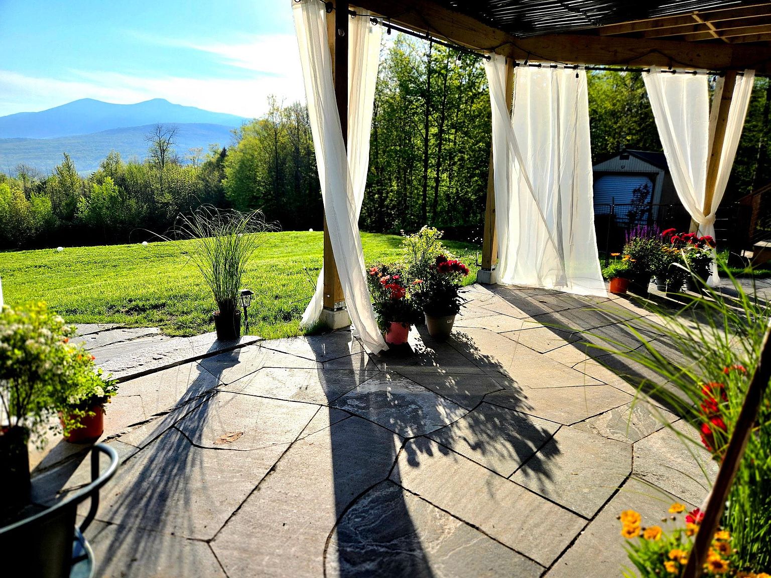 This inviting stone-paved patio features a covered pergola draped with elegant white curtains, creating a soft, breezy atmosphere. The space overlooks a lush green lawn that transitions into a scenic mountain vista, offering a peaceful and picturesque outdoor living area. Potted flowers and ornamental grasses add vibrant color and texture to the stone terrace, enhancing the serene, resort-like feel of the property.