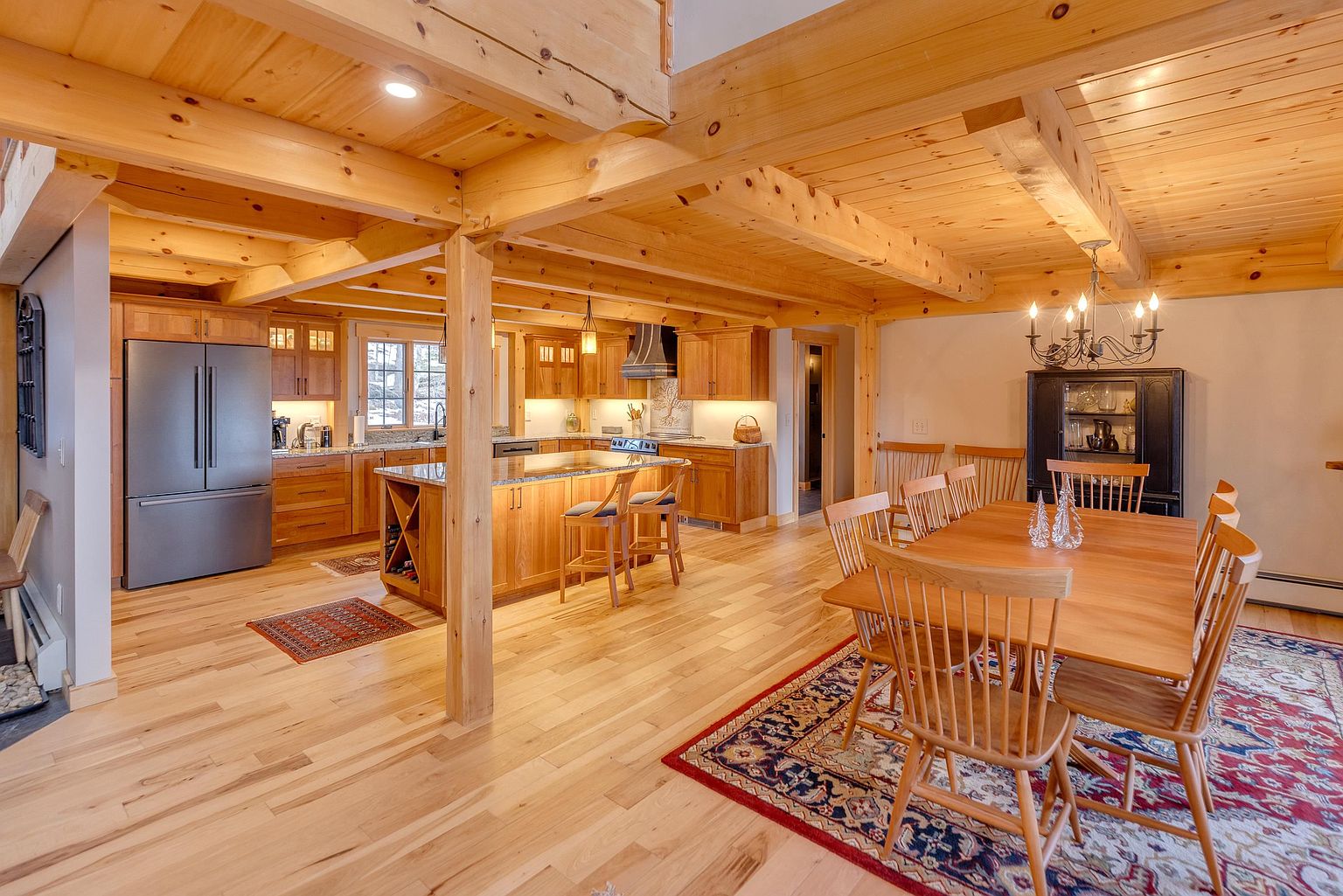 This open-concept space features a warm, rustic aesthetic with exposed light wood ceiling beams and matching cabinetry throughout. The kitchen is equipped with stainless steel appliances and a central island with bar seating, while the adjacent dining area is anchored by a wooden table and a traditional chandelier. The perspective captures the seamless flow between the culinary workspace and the dining room, highlighting the cohesive wood finishes and natural light.