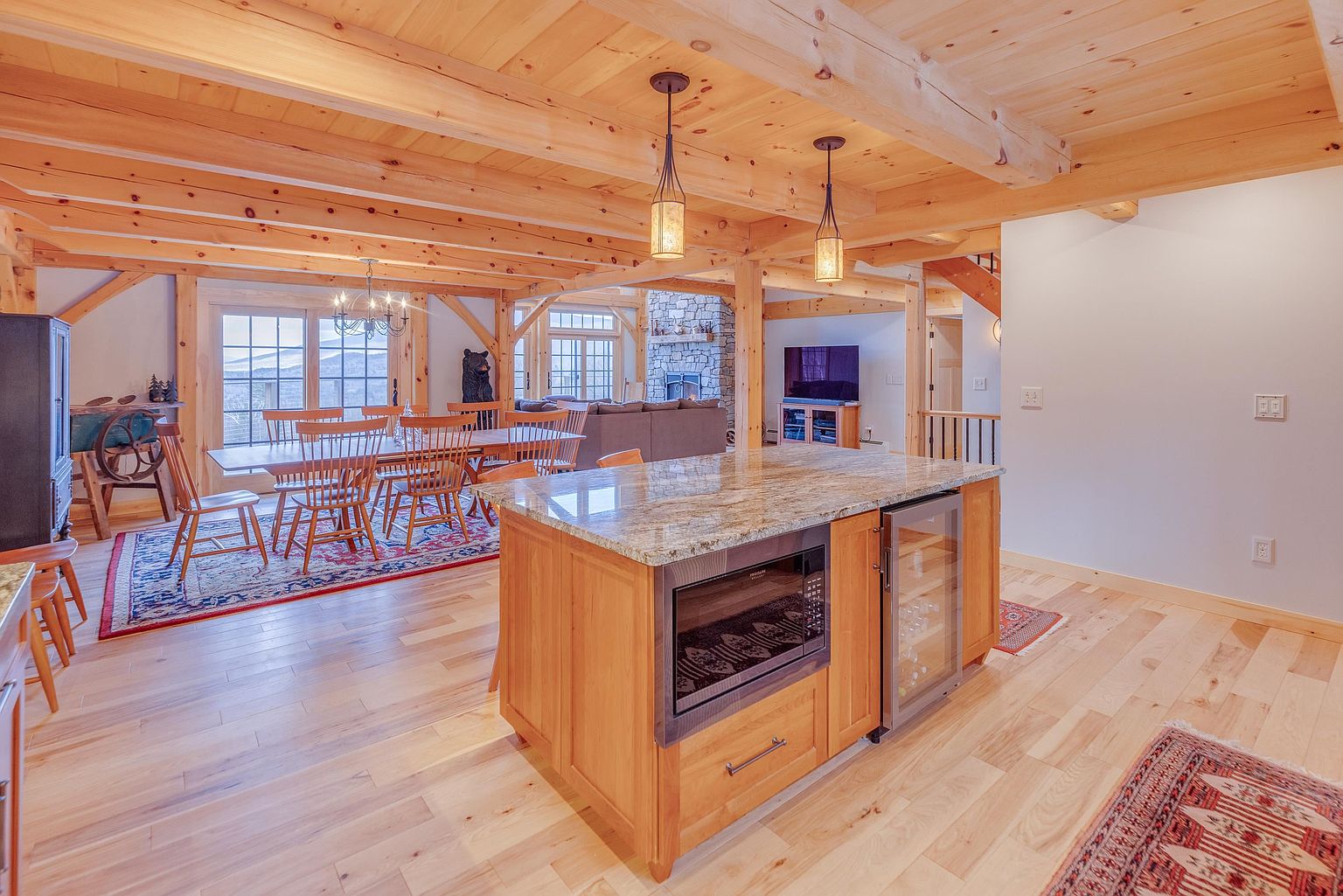 This open-concept kitchen and dining area features exposed timber beams and light wood flooring, creating a warm, rustic atmosphere. A central kitchen island with a stone countertop houses a built-in microwave and a glass-front beverage cooler, while the adjacent dining space is anchored by a large wooden table and a patterned area rug. The perspective looks from the kitchen island toward the dining area and a living space with a stone-clad chimney breast in the background.