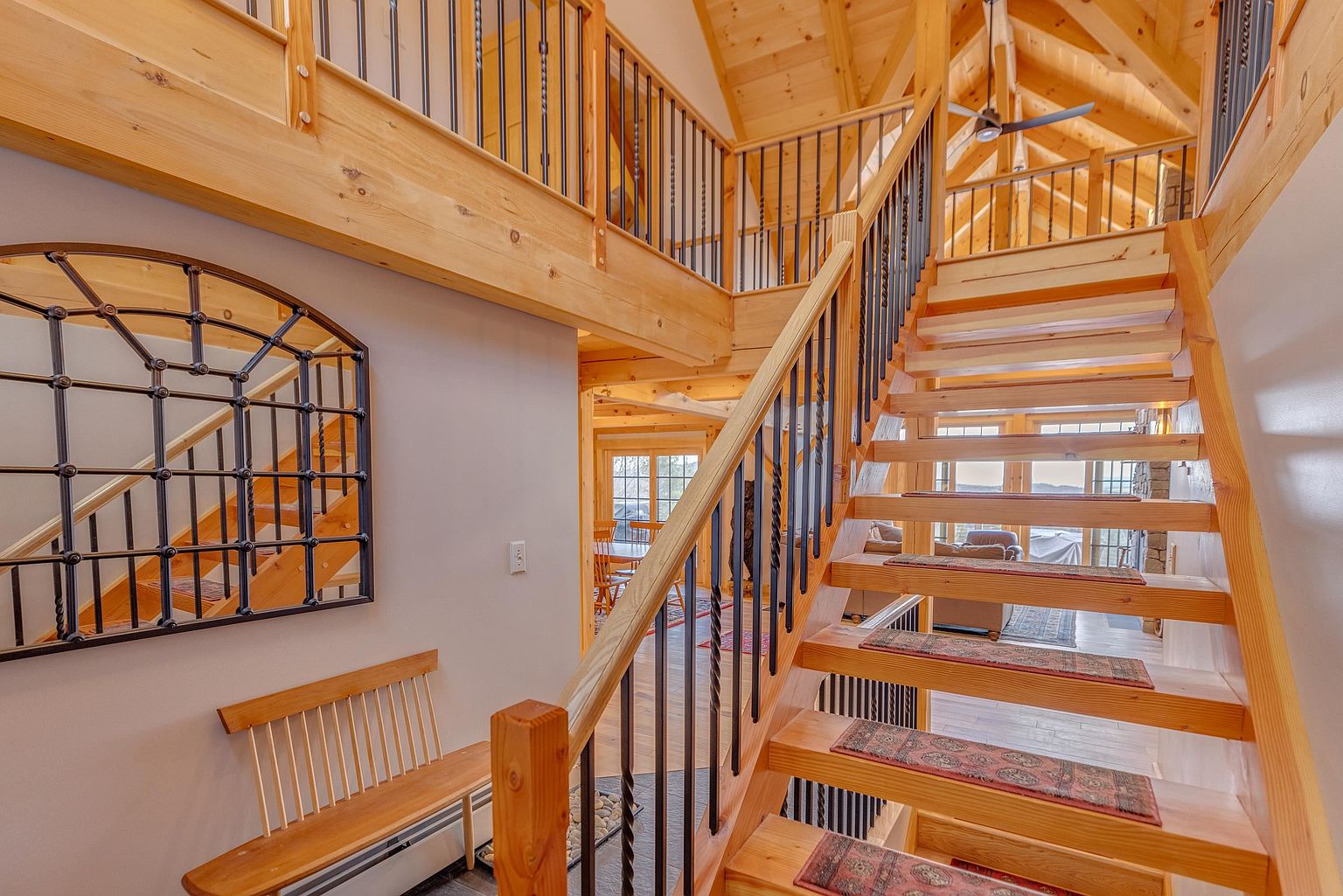 This interior view captures a grand, open-concept staircase featuring light wood treads with patterned runners and black metal balusters. To the left, a wooden bench sits beneath a large, arched decorative mirror, while the open design allows for a clear line of sight through to the living area and dining space beyond. The vaulted ceiling with exposed timber beams adds a rustic, airy feel to the entryway and landing area.