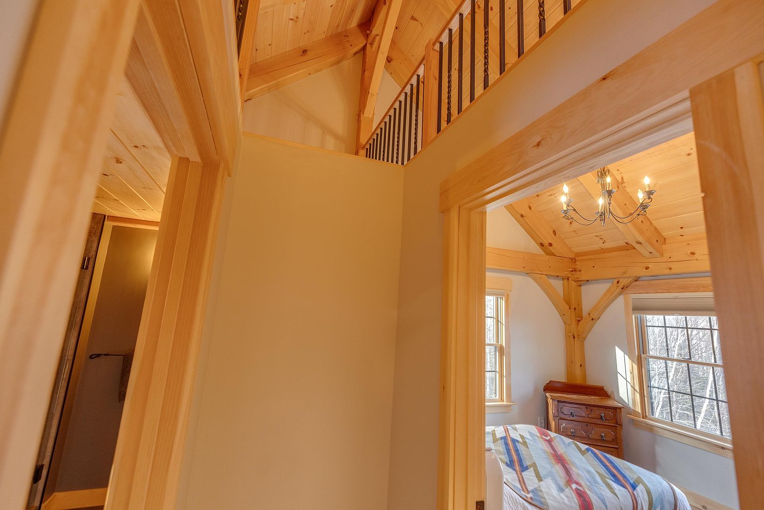 This perspective captures a transition space featuring a light-toned wood staircase with a metal railing that overlooks a bedroom below. The view through the doorway reveals a cozy bedroom with exposed timber beams, a chandelier, and a bed dressed in a patterned quilt. The overall aesthetic is rustic and warm, emphasizing the architectural character of the timber framing.
