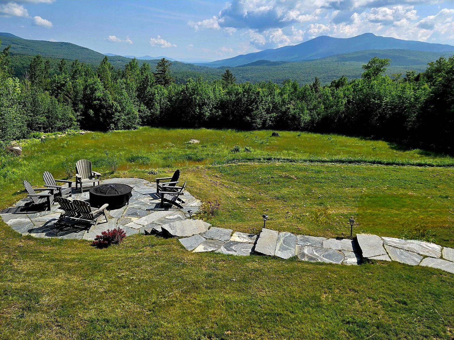 This elevated perspective captures a stone-paved patio area featuring a circular fire pit surrounded by Adirondack chairs, set against a backdrop of lush green fields and rolling mountains. A stone walkway leads from the patio across the lawn, emphasizing the expansive outdoor living space and scenic natural views. The scene conveys a serene, private atmosphere ideal for relaxation and enjoying the landscape.