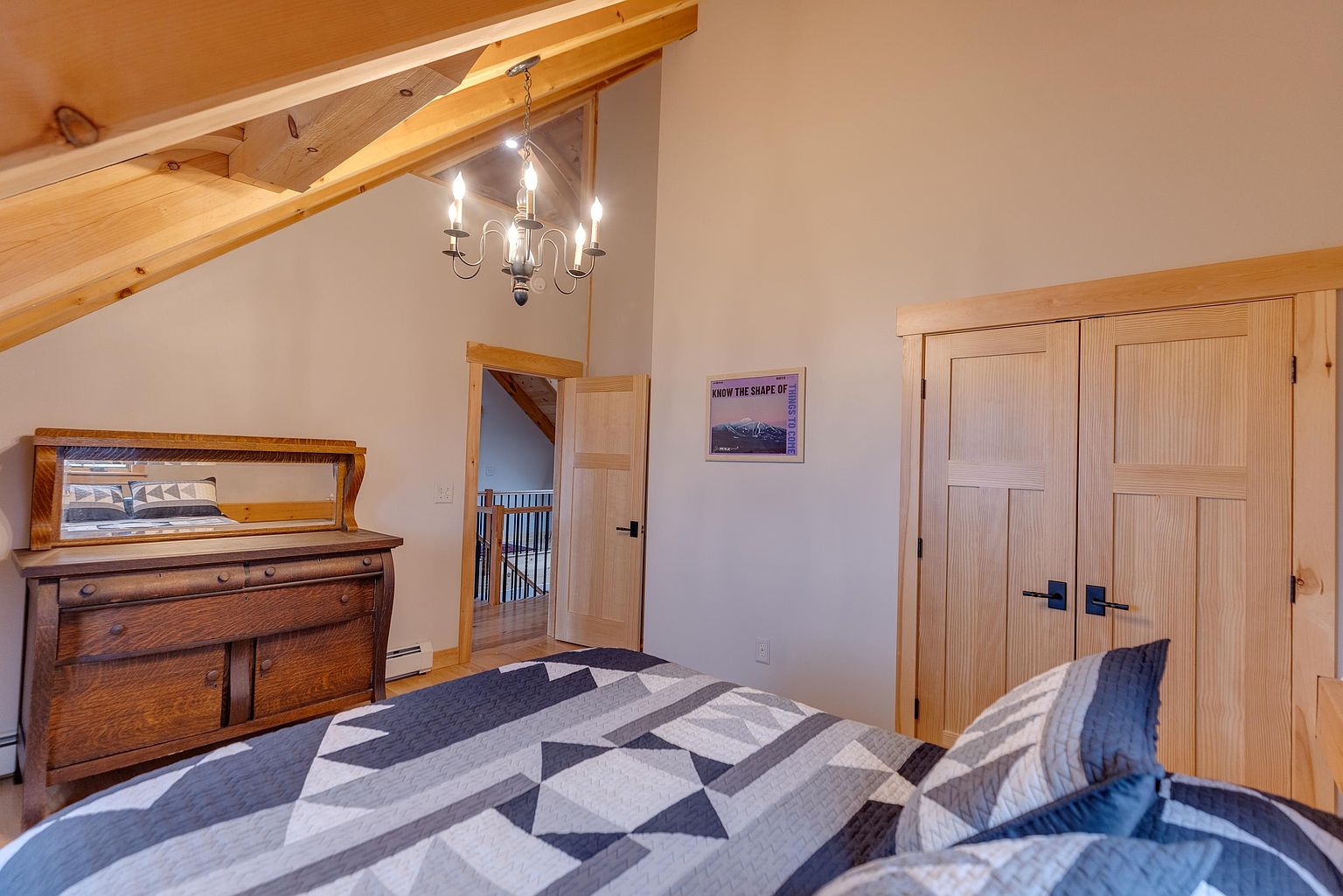 This cozy bedroom features a vaulted ceiling with exposed light wood beams and a rustic chandelier, creating a warm, cabin-like atmosphere. The room is furnished with a patterned quilt on the bed, a vintage-style wooden dresser with an integrated mirror, and a double-door closet with light wood paneling. A framed mountain-themed print hangs on the wall, and an open doorway leads to a hallway with a wooden staircase railing visible in the background.