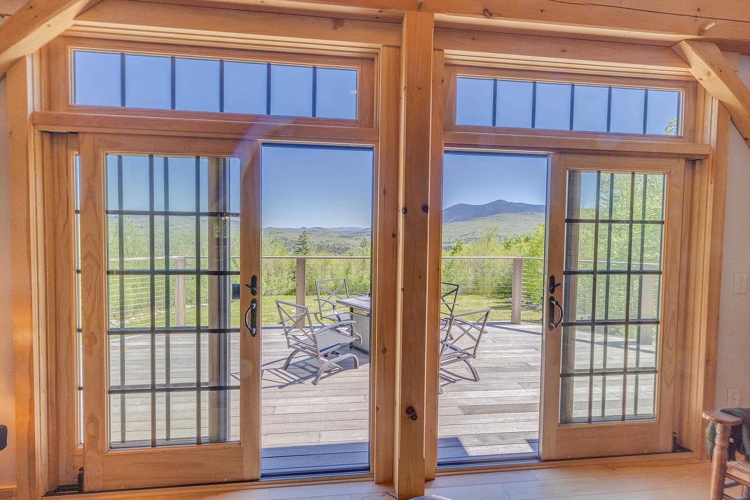 This interior view captures a pair of large, light-wood framed sliding glass doors that open onto a spacious wooden deck. The doors feature a grid-patterned design and are topped with transom windows, allowing for an abundance of natural light and a clear view of the surrounding forest and distant mountains. The perspective is from inside a room looking out, emphasizing the seamless transition between the indoor living space and the outdoor deck area.