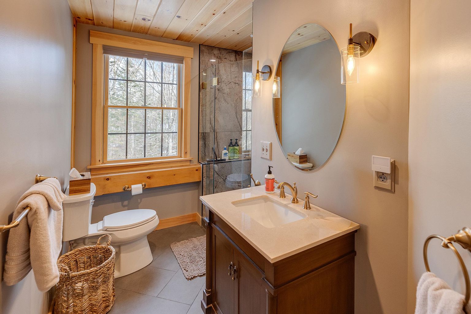 This bathroom features a rustic-modern aesthetic with a wood-paneled ceiling and a vanity topped with a light-colored stone counter. A large oval mirror is flanked by two wall-mounted light fixtures, while a window with wood trim provides natural light and a view of the outdoors. The space includes a toilet, a woven basket for storage, and a glass-enclosed shower area with dark stone-look wall tiles.