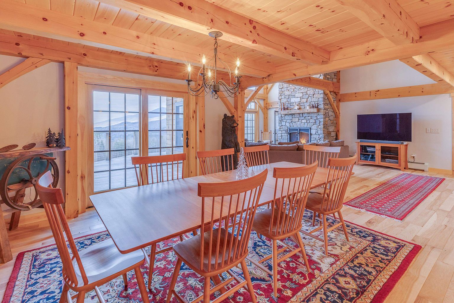 This open-concept dining area features a large wooden table surrounded by spindle-back chairs, set atop a vibrant, intricately patterned rug. The space is defined by exposed timber beams and light wood flooring, creating a warm, rustic atmosphere that flows into an adjacent living area with a stone-clad chimney breast. Large sliding glass doors provide a view of the outdoors, while a classic chandelier hangs above the table to anchor the room.