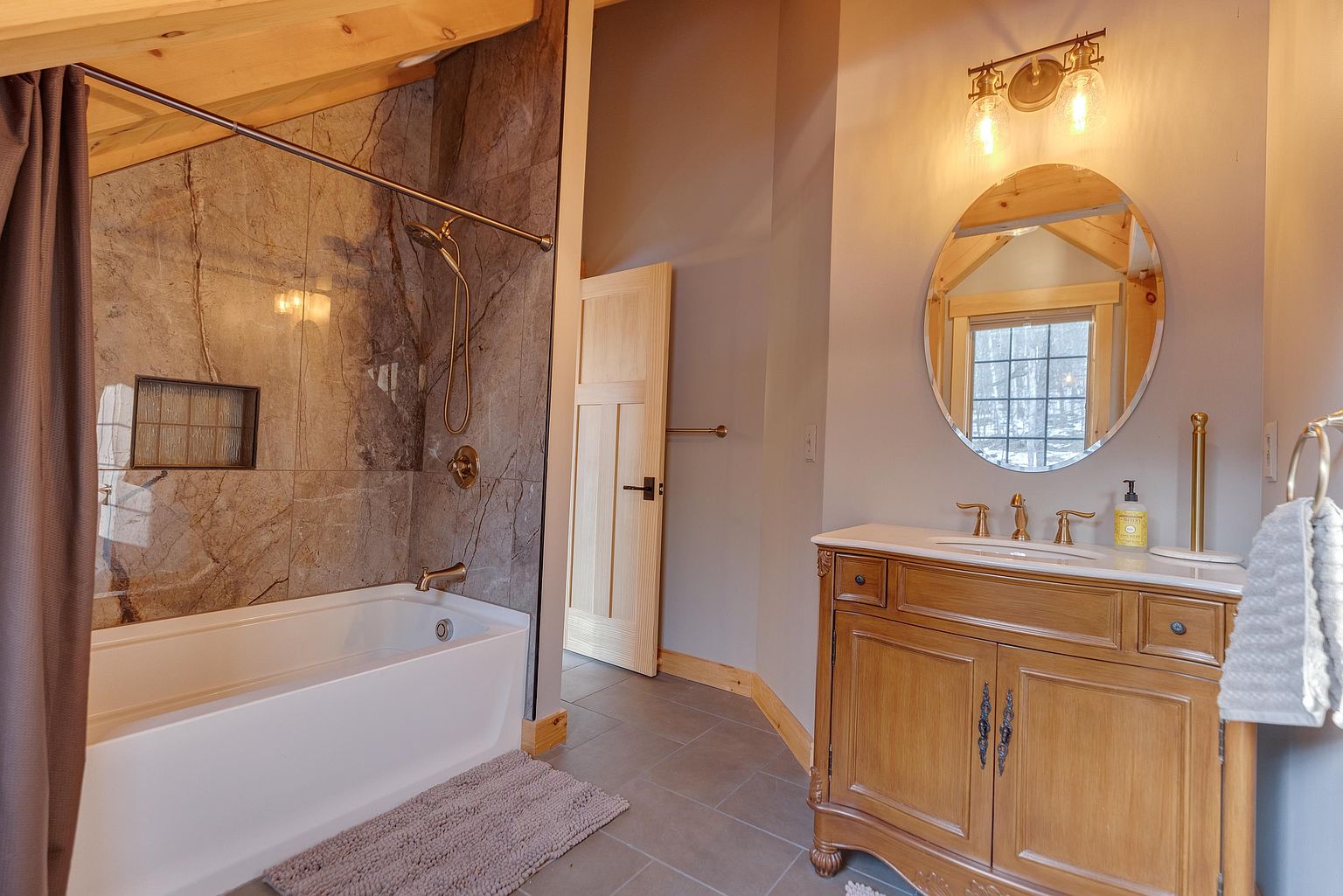 This bathroom features a rustic aesthetic with a light wood vanity topped with a stone counter and a round mirror. To the left, a bathtub is enclosed by a shower curtain and surrounded by large, textured stone-look wall tiles. The room includes a wooden door, neutral wall tones, and warm lighting, creating a cozy and inviting atmosphere.