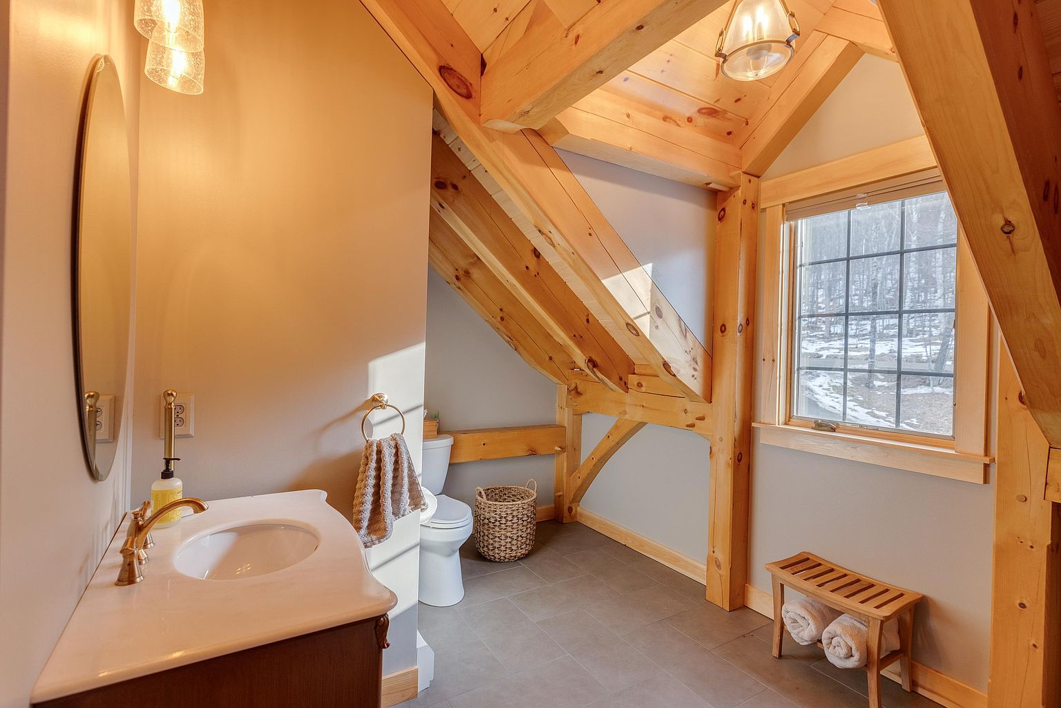 This rustic-style bathroom features exposed timber beams and a vaulted ceiling, creating a warm and cozy atmosphere. The space includes a vanity with a stone countertop, a toilet tucked under the sloped roofline, and a wooden bench holding rolled towels. A window provides natural light, highlighting the neutral wall tones and dark tile flooring.