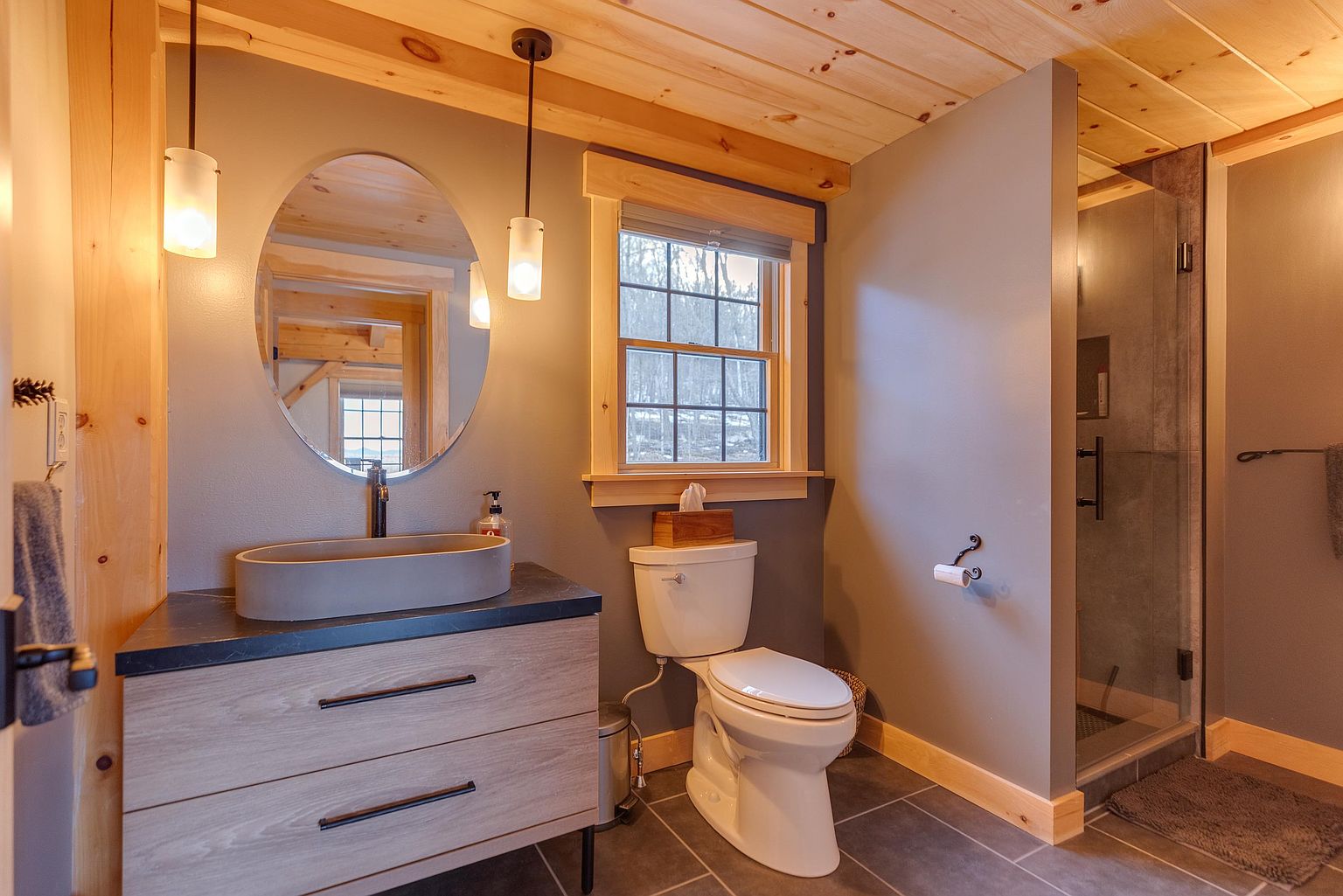 This modern bathroom features a rustic-chic aesthetic with light wood cabinetry, a dark stone countertop, and a sleek oval vessel sink. The space is accented by a wood-paneled ceiling, a glass-enclosed shower, and a window that brings in natural light. The neutral color palette and clean lines create a warm, inviting atmosphere.