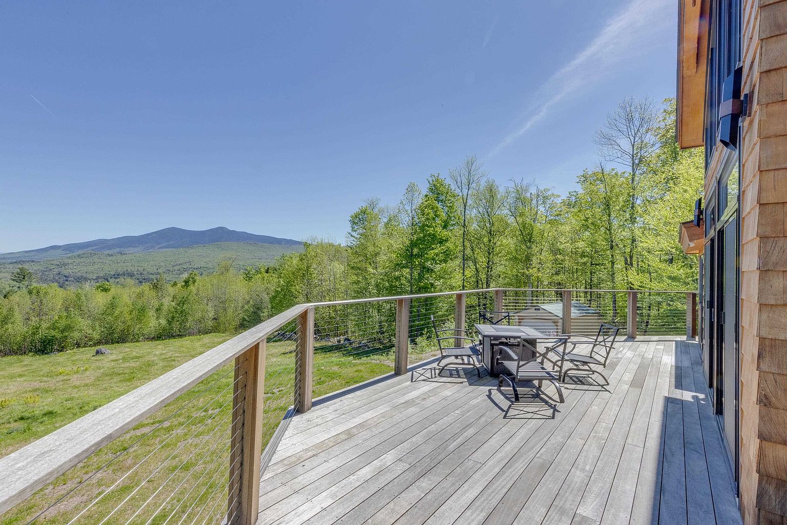 This elevated wooden deck offers a panoramic view of a lush, green mountain landscape under a clear blue sky. The space is furnished with a small outdoor table and chairs, featuring a modern cable railing system that maintains an unobstructed sightline. The perspective captures the expansive outdoor living area as it transitions from the home's cedar-shingled exterior toward the natural scenery beyond.