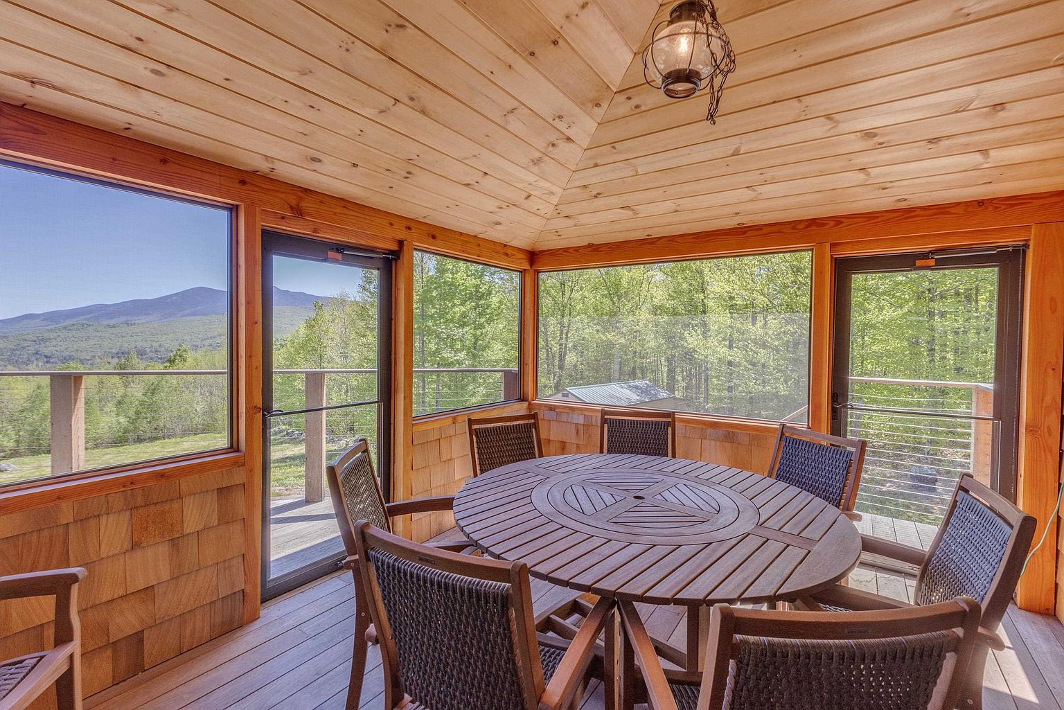 This inviting screened-in porch features a vaulted wood-paneled ceiling and a central round wooden dining table surrounded by six chairs. Large screened windows provide expansive, unobstructed views of the surrounding forest and distant mountains, creating a seamless connection to the outdoors. The space is finished with warm wood walls and flooring, offering a cozy yet airy atmosphere perfect for dining or relaxation.
