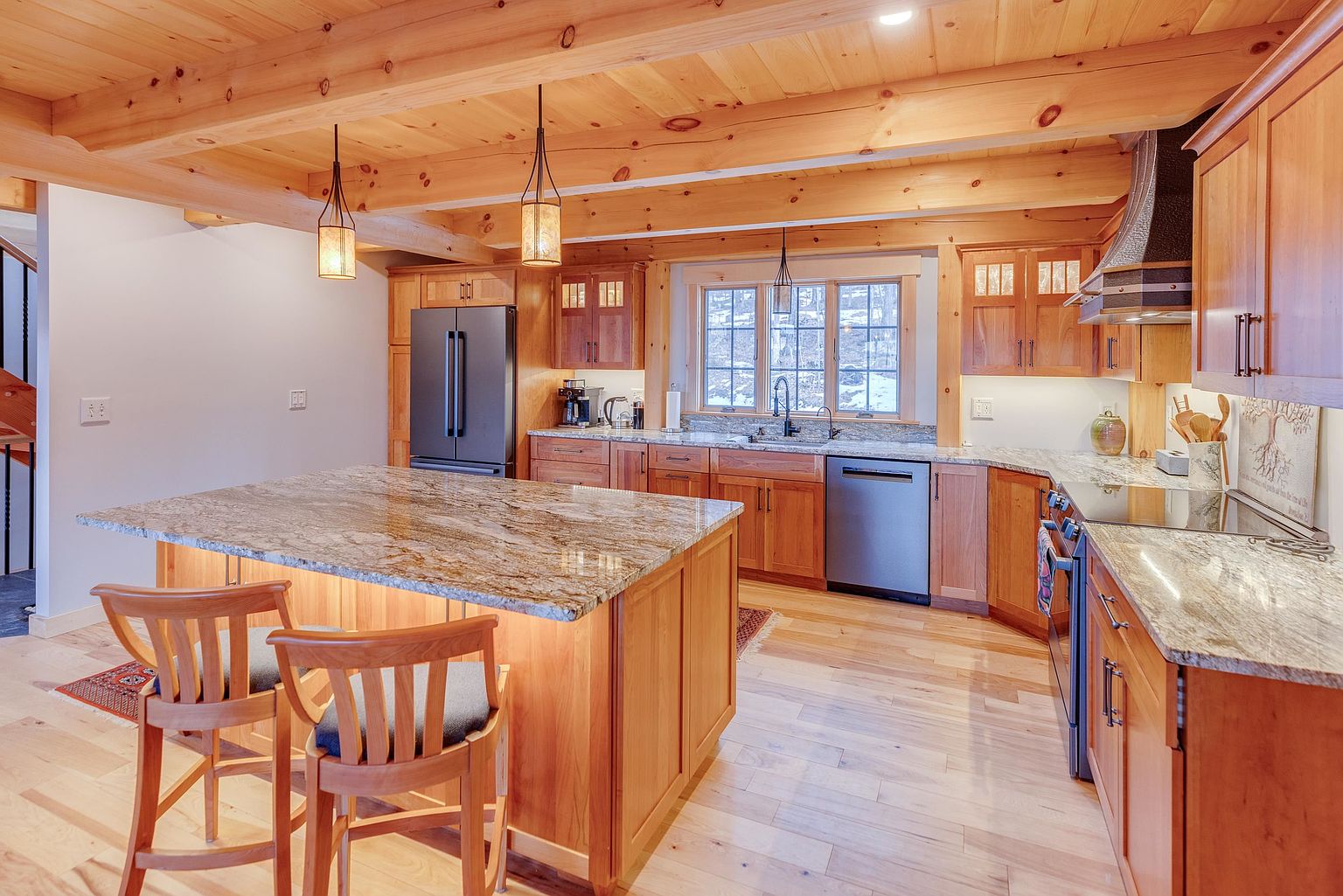 This warm and inviting kitchen features natural wood cabinetry, exposed ceiling beams, and a large central island topped with patterned stone. Stainless steel appliances, including a refrigerator and dishwasher, are integrated into the layout, while pendant lights hang above the island to provide focused illumination. The space is brightened by a window over the sink, creating a cozy, rustic-inspired atmosphere perfect for family gatherings.
