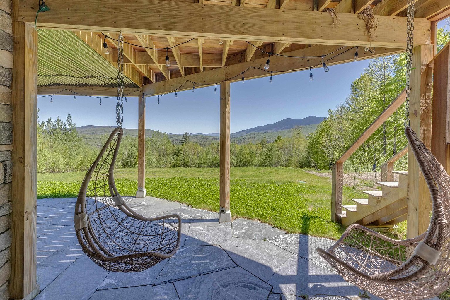 This covered patio features a natural stone floor and two hanging wicker chairs, creating a relaxing outdoor living space. The structure is supported by wooden posts and beams, with string lights providing ambient illumination. Beyond the patio, a wooden staircase leads to the yard, offering an expansive view of the surrounding forest and distant mountains.