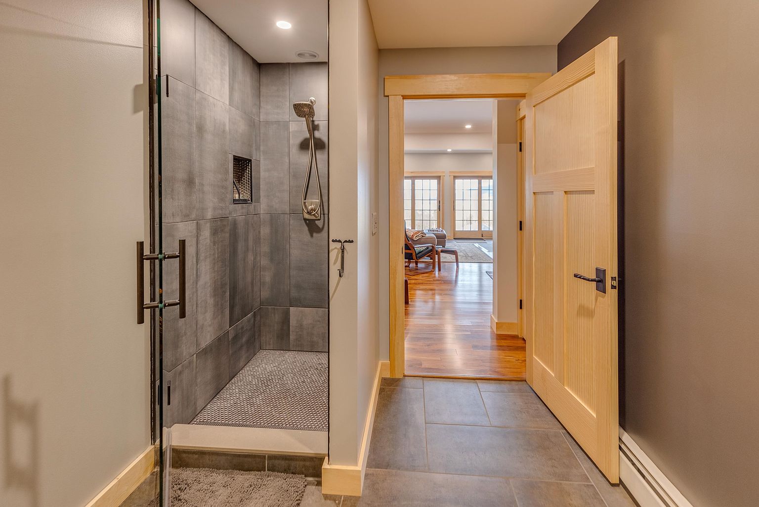 This interior view captures a modern bathroom featuring a walk-in shower with large-format grey tiles and a glass door. A light wood door with a dark handle leads into an adjacent living space with hardwood floors and large windows. The neutral color palette and clean lines create a contemporary and functional aesthetic.
