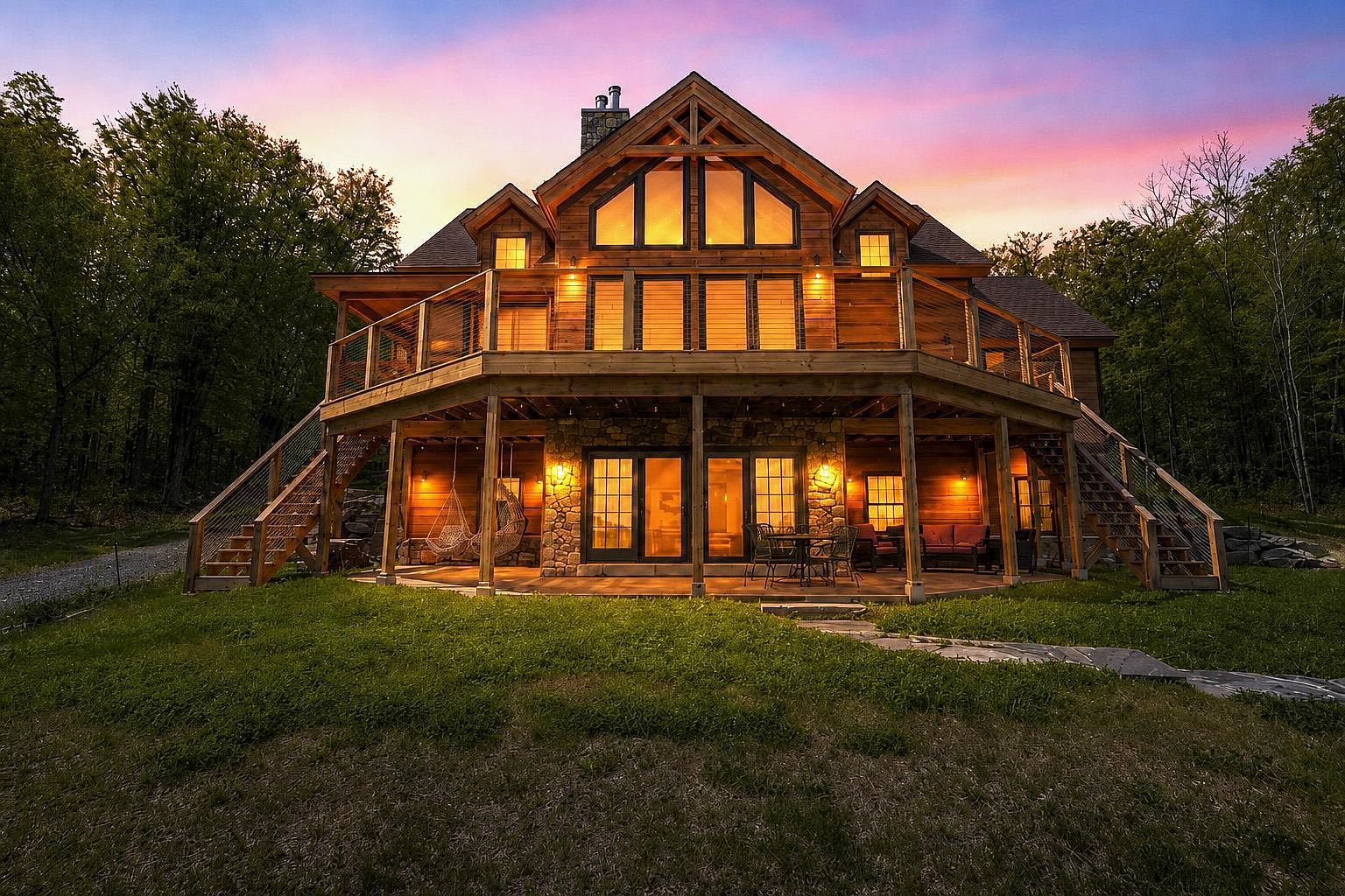 This stunning timber-frame home is captured at dusk, showcasing a multi-level design with a expansive wooden deck and a stone-clad lower patio. The warm glow from the interior lighting and exterior sconces highlights the natural wood siding and stone accents, creating an inviting and cozy atmosphere. The property is nestled against a lush forest backdrop, with a vibrant sunset sky providing a dramatic and picturesque setting.