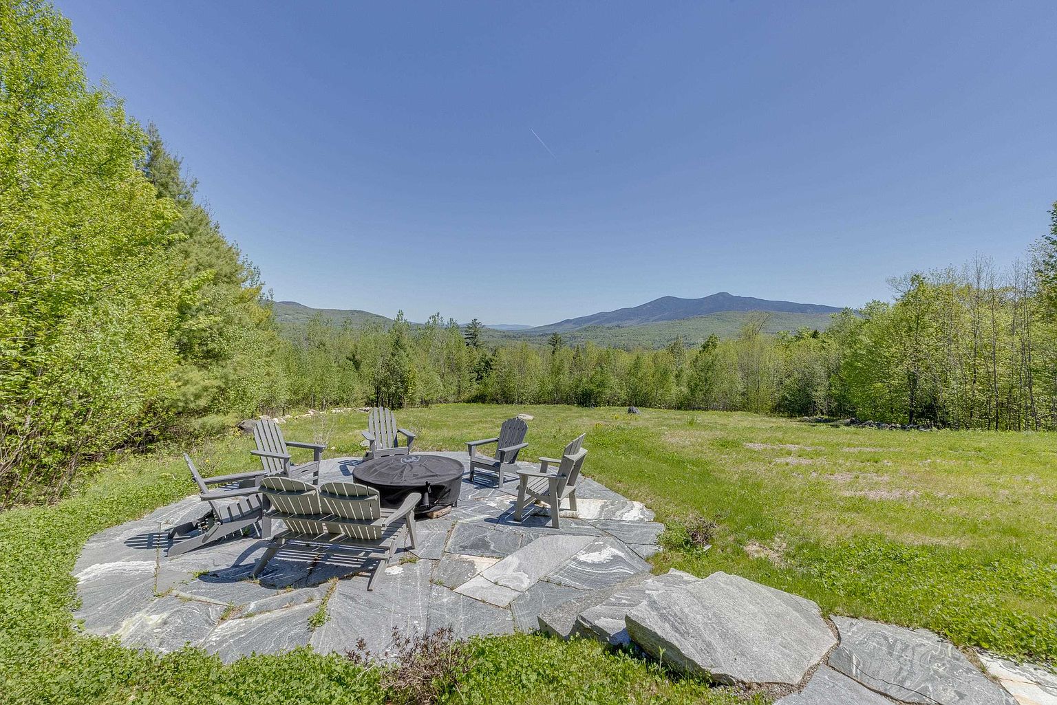 This outdoor space features a natural stone patio arranged with several Adirondack chairs surrounding a central fire pit. The patio is set against a backdrop of a sprawling grassy lawn that transitions into a dense forest, with majestic mountain ranges visible in the distance under a clear blue sky. The perspective is a wide-angle shot from a slightly elevated position, capturing the serene and expansive nature of the property.