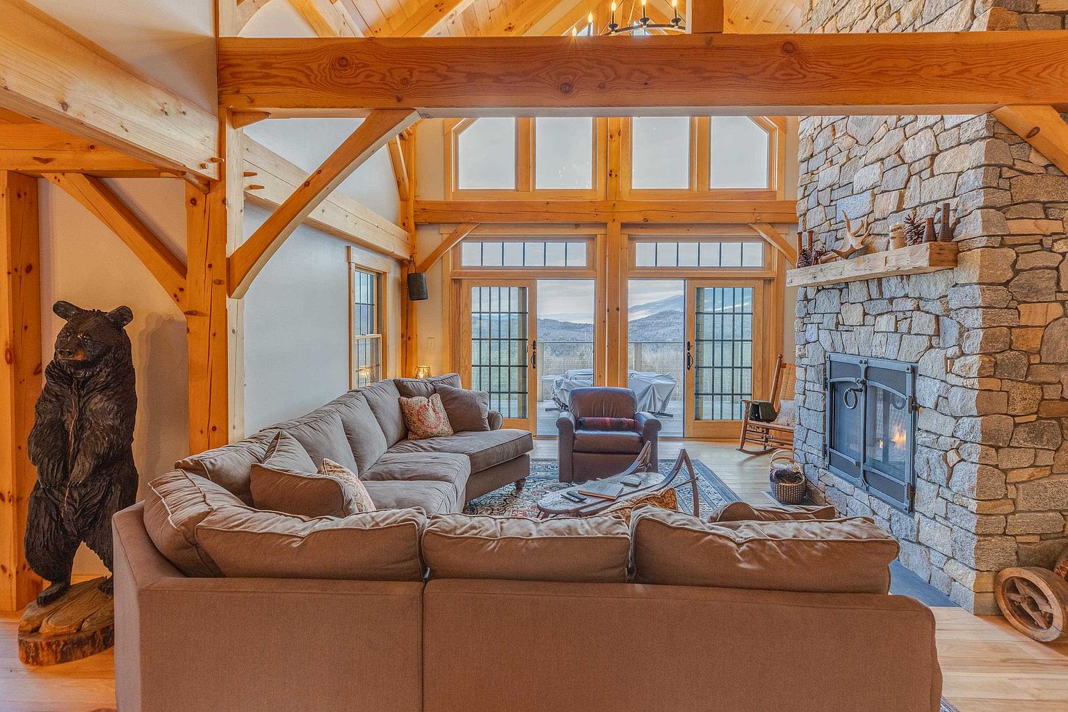 This spacious living room features a rustic timber-frame aesthetic with exposed beams and a prominent stone-clad chimney breast. A large sectional sofa and a leather armchair are arranged around a central coffee table, creating a cozy seating area that faces a set of tall glass doors leading to a deck. The room is accented by a decorative bear sculpture and offers a warm, inviting atmosphere with views of the surrounding landscape.