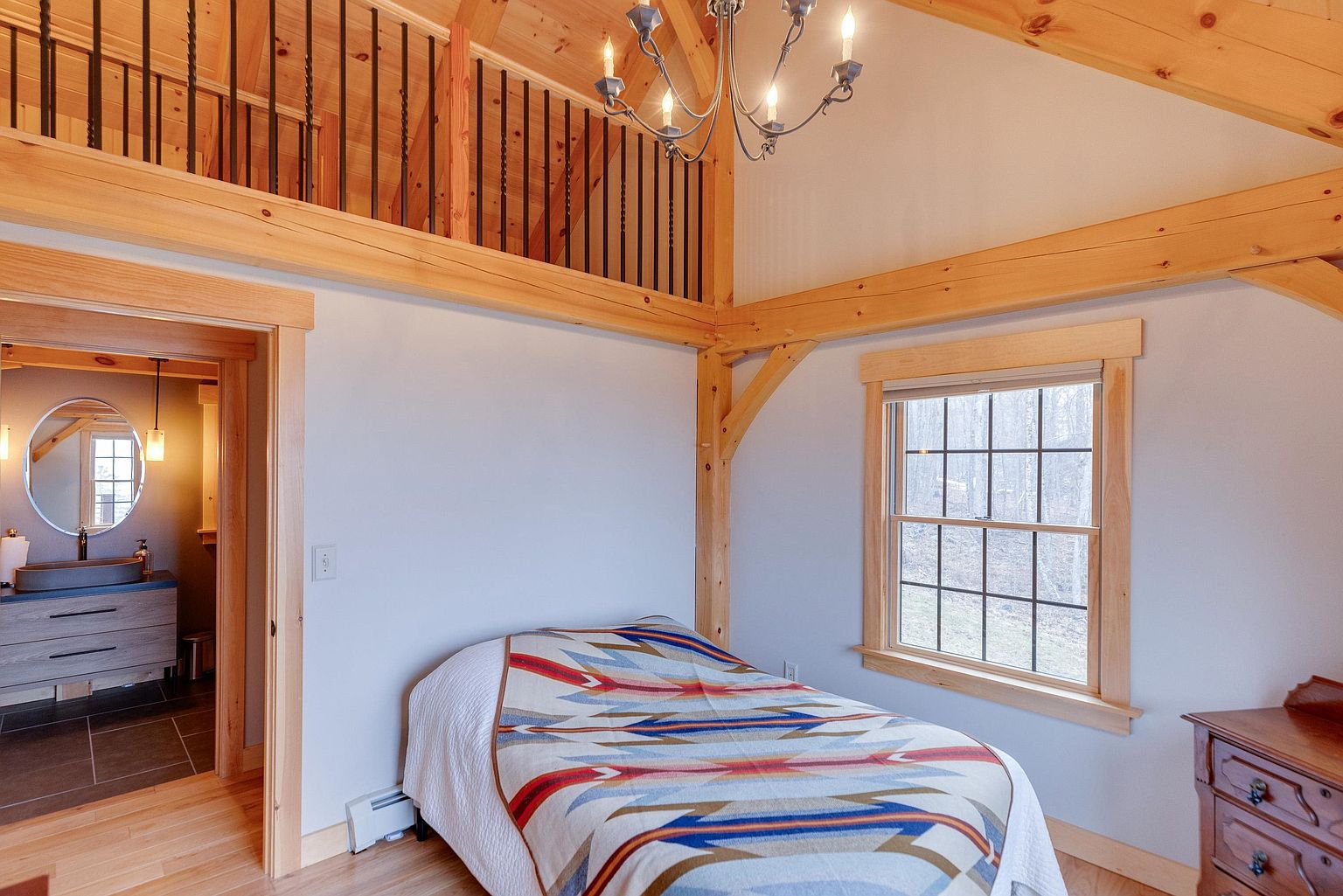 This cozy bedroom features a rustic aesthetic with exposed timber beams and a vaulted ceiling, creating an airy and inviting atmosphere. A bed with a vibrant, patterned quilt serves as the focal point, positioned beneath a classic chandelier and adjacent to a window that provides natural light. The room also offers a direct view into an adjoining bathroom, highlighting a seamless transition between living and personal spaces.