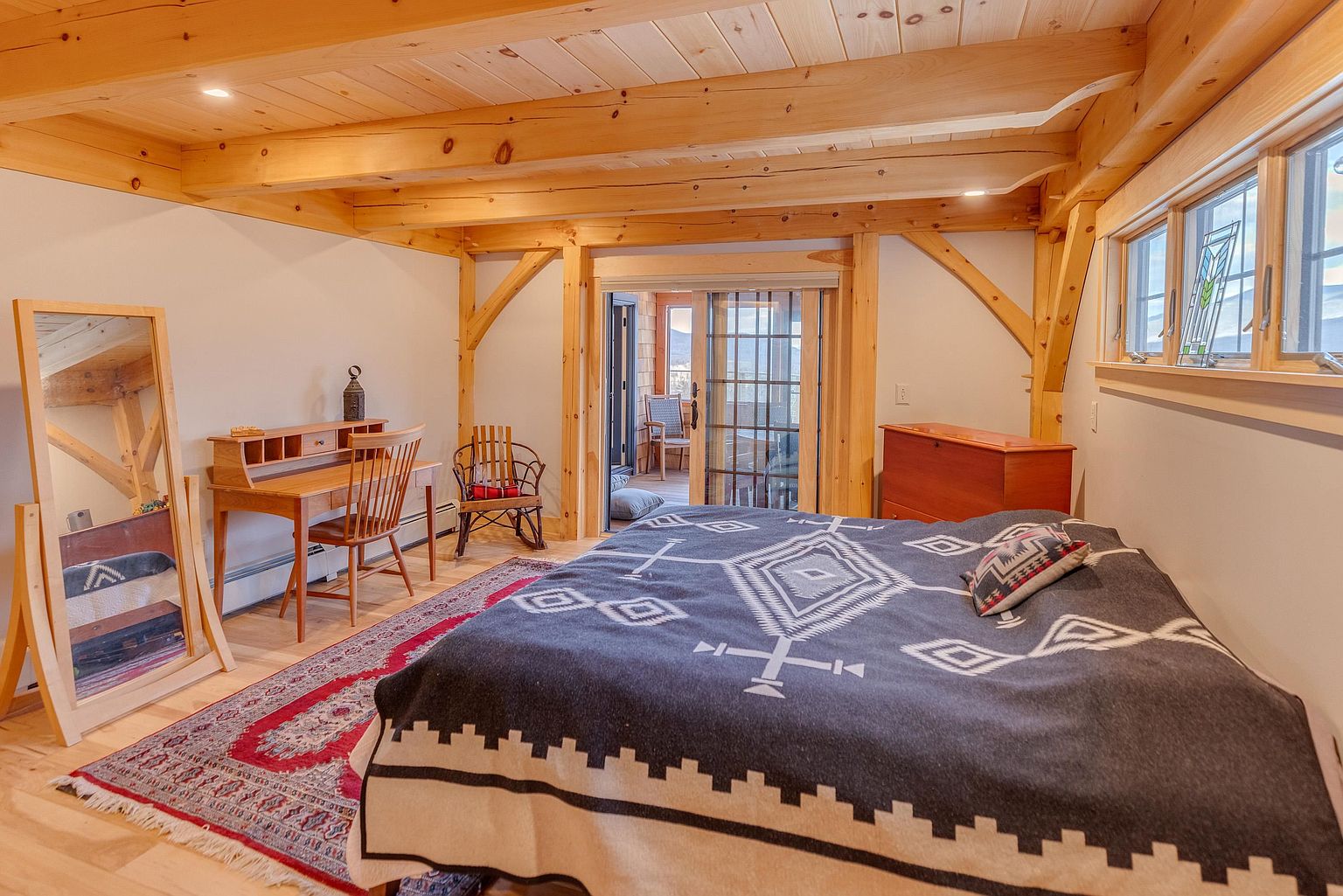 This cozy bedroom features exposed timber beams and a rustic aesthetic, centered around a bed dressed in a patterned textile. To the left, a wooden desk and chair are positioned near a full-length floor mirror, while a sliding glass door leads to an outdoor deck. The room is anchored by a traditional patterned rug, creating a warm and inviting atmosphere.