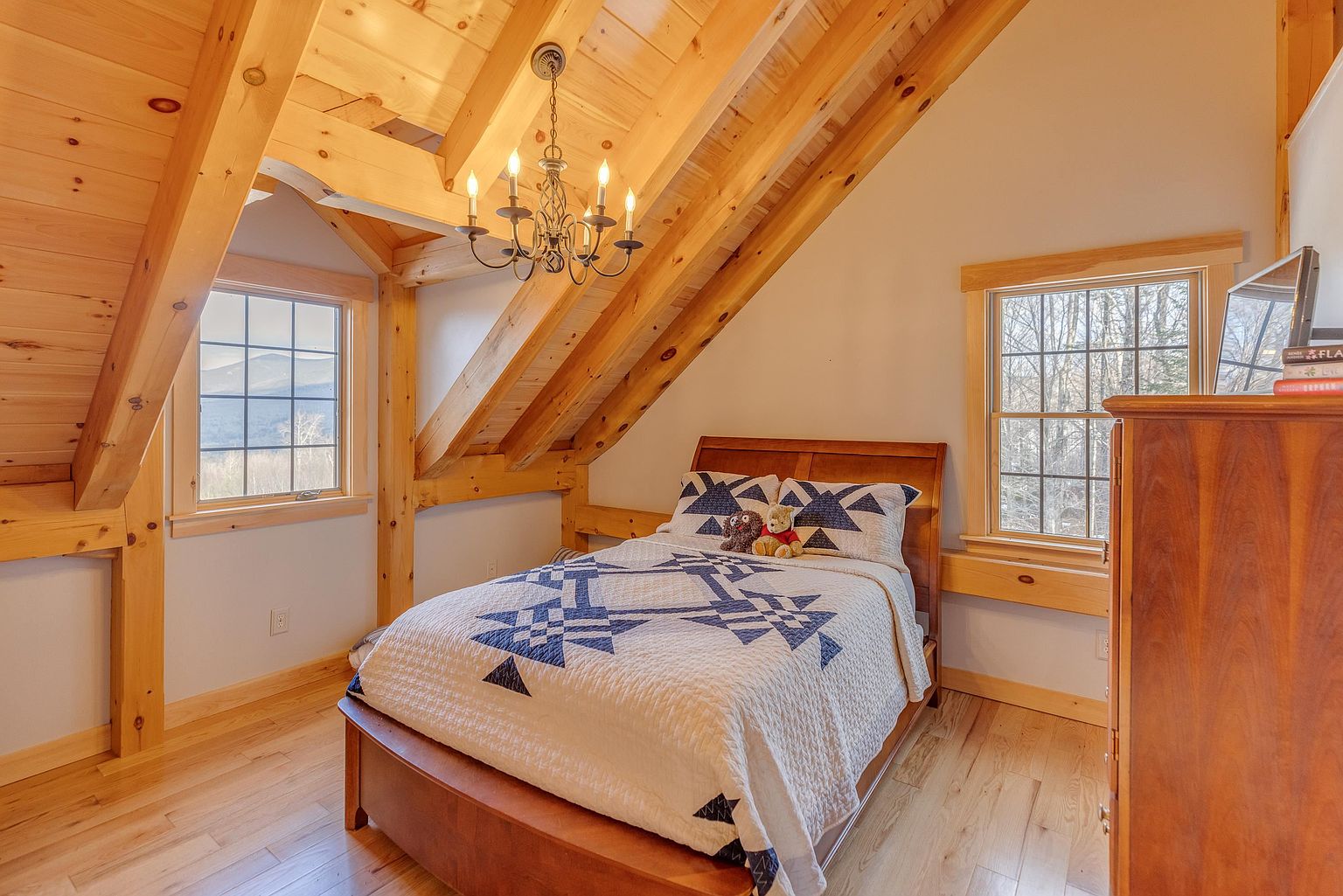 This cozy bedroom features a rustic aesthetic with exposed light wood beams and vaulted ceilings. A bed with a blue and white patterned quilt sits centrally in the room, flanked by two windows that provide natural light and views of the outdoors. The warm wood flooring and timber framing create a charming, cabin-like atmosphere.