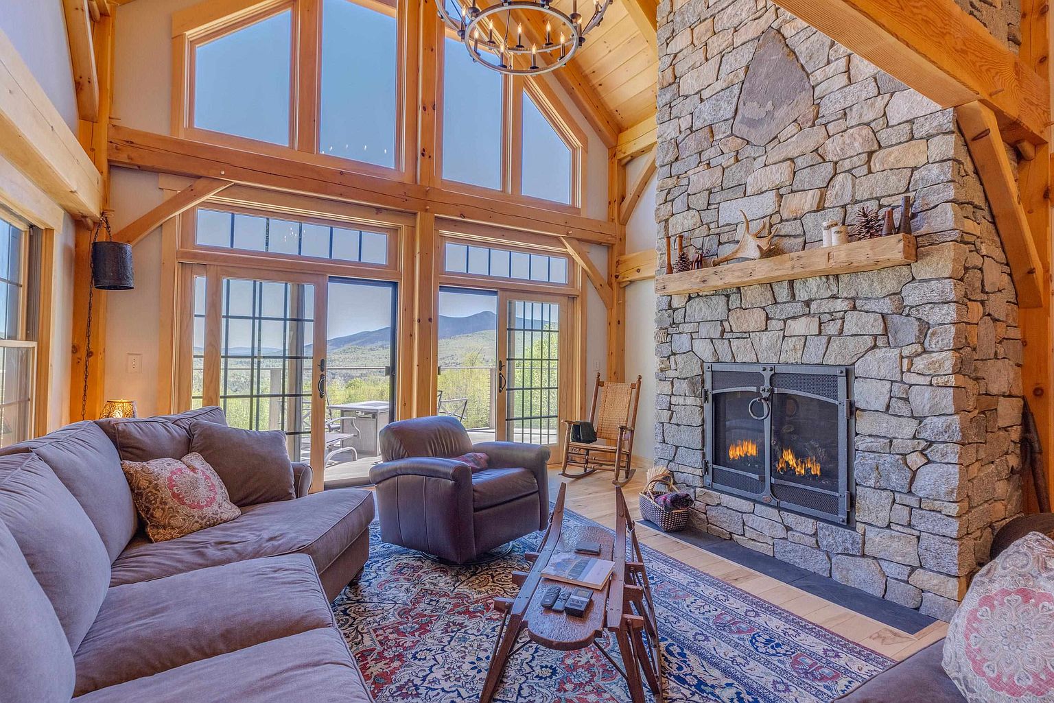 This rustic living room features a prominent stone-clad chimney breast and a large, exposed timber frame structure. The space is anchored by a plush sofa and a leather armchair arranged on a patterned area rug, with tall windows providing expansive views of the surrounding landscape. A wooden coffee table and a rocking chair add to the cozy, mountain-lodge aesthetic of the room.