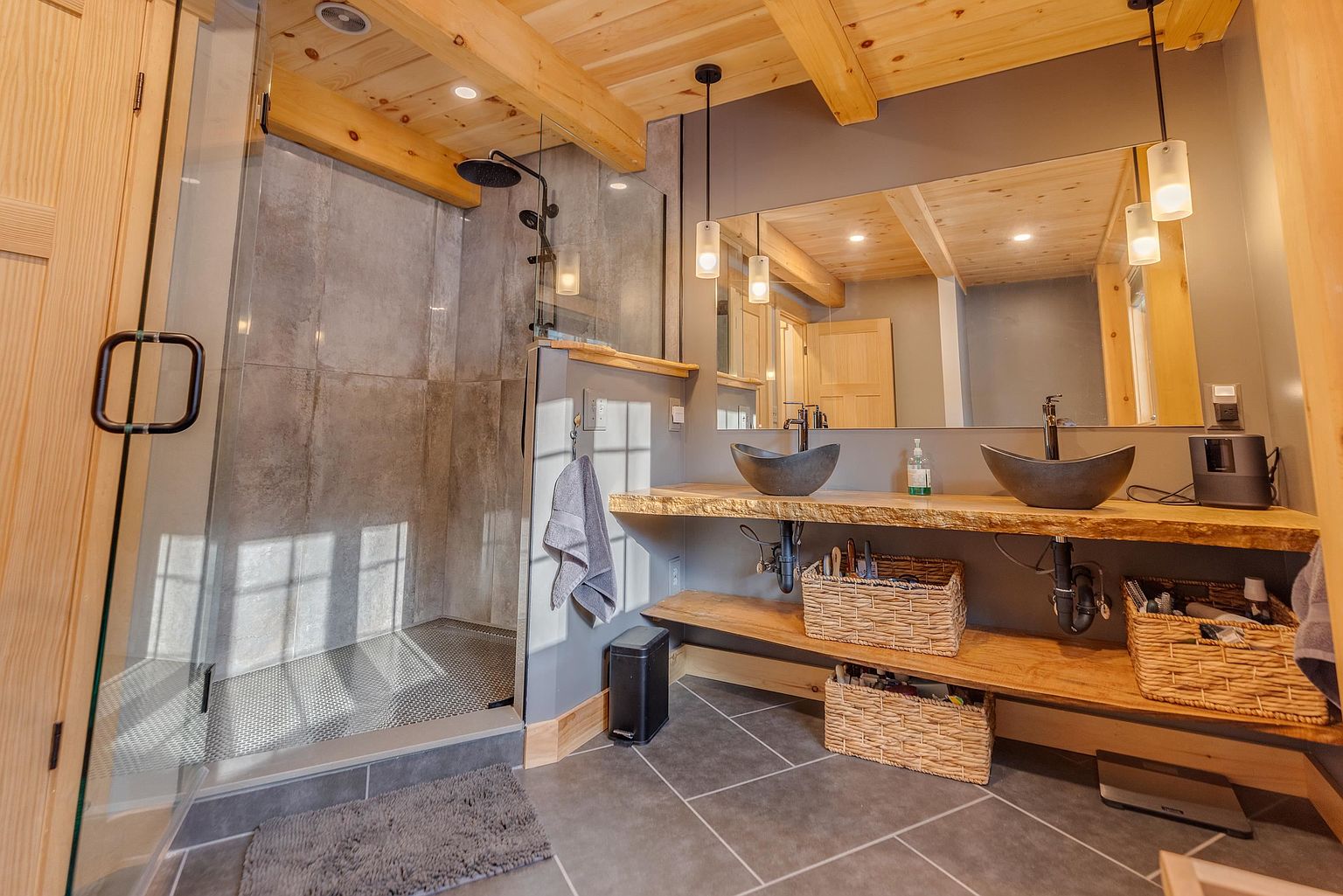 This rustic-modern bathroom features a dual-sink vanity with stone vessel sinks resting on a live-edge wood slab countertop. A large walk-in shower with a glass enclosure and gray tile walls sits adjacent to the vanity, while exposed wood beams on the ceiling add warmth to the space. The room is accented by dark gray floor tiles and woven storage baskets, creating a cohesive and earthy aesthetic.