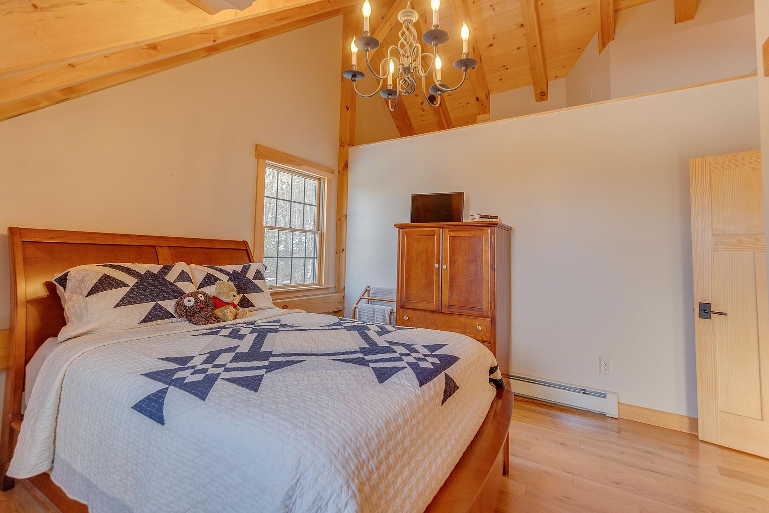 This cozy bedroom features a vaulted ceiling with exposed light wood beams and a rustic chandelier, creating a warm and inviting atmosphere. The room is furnished with a bed dressed in a geometric-patterned quilt and a wooden armoire that serves as a focal point. A window to the left allows natural light to brighten the space, which is finished with light-toned wood flooring.