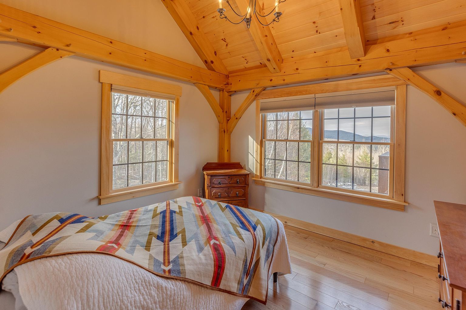 This cozy bedroom features exposed timber beams and a vaulted ceiling, creating a rustic and inviting atmosphere. A bed with a patterned, multi-colored blanket sits centrally in the room, flanked by two windows that offer natural light and views of the surrounding landscape. A small wooden dresser is positioned in the corner, and the light wood flooring adds to the warm, cabin-like aesthetic of the space.