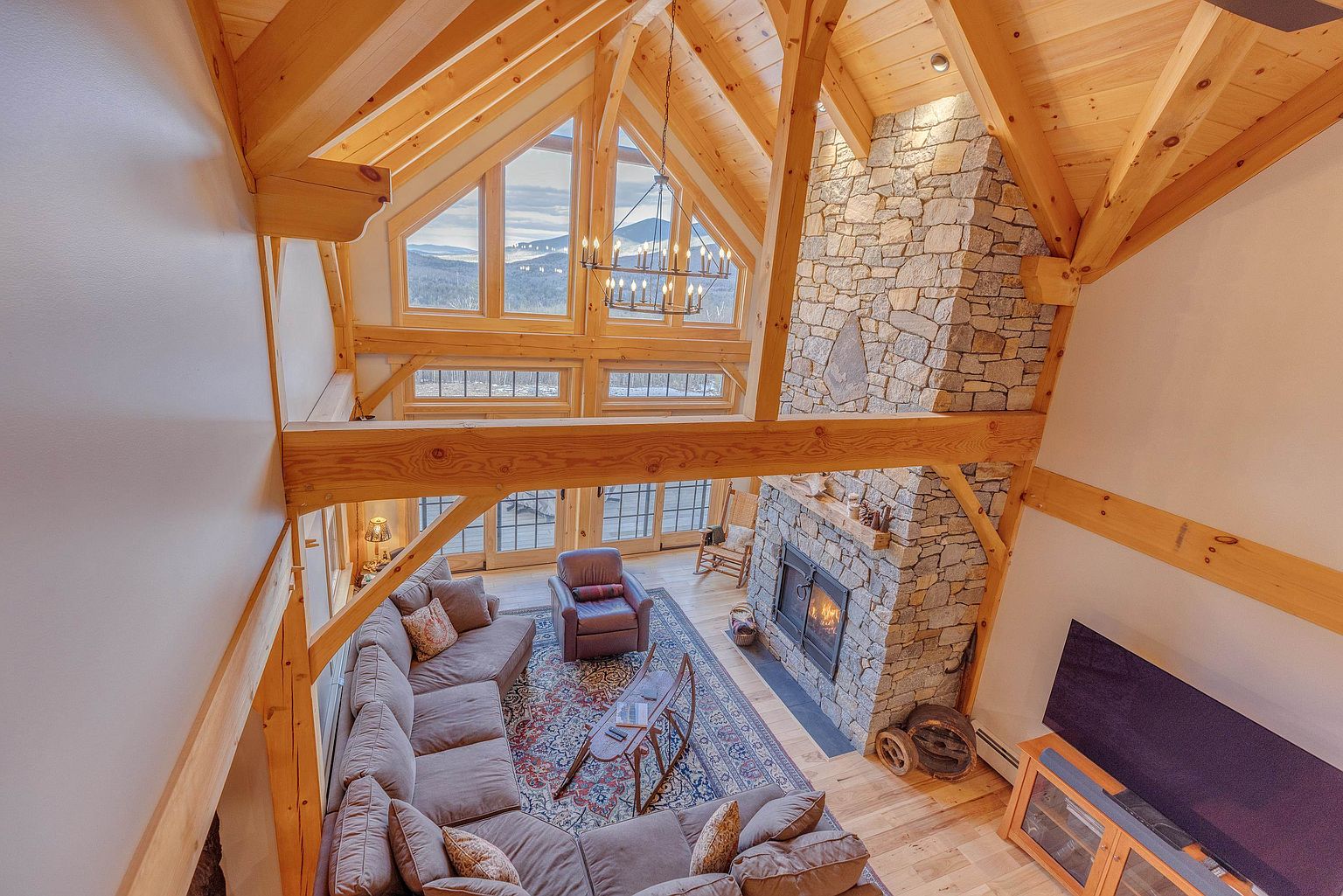 This high-angle shot captures a spacious, rustic living room characterized by exposed timber framing and a vaulted ceiling. A prominent stone-clad chimney breast serves as the focal point, complemented by a large sectional sofa, a patterned area rug, and expansive windows that frame a scenic mountain view. The warm, natural wood tones and open-concept design create an inviting and airy atmosphere.