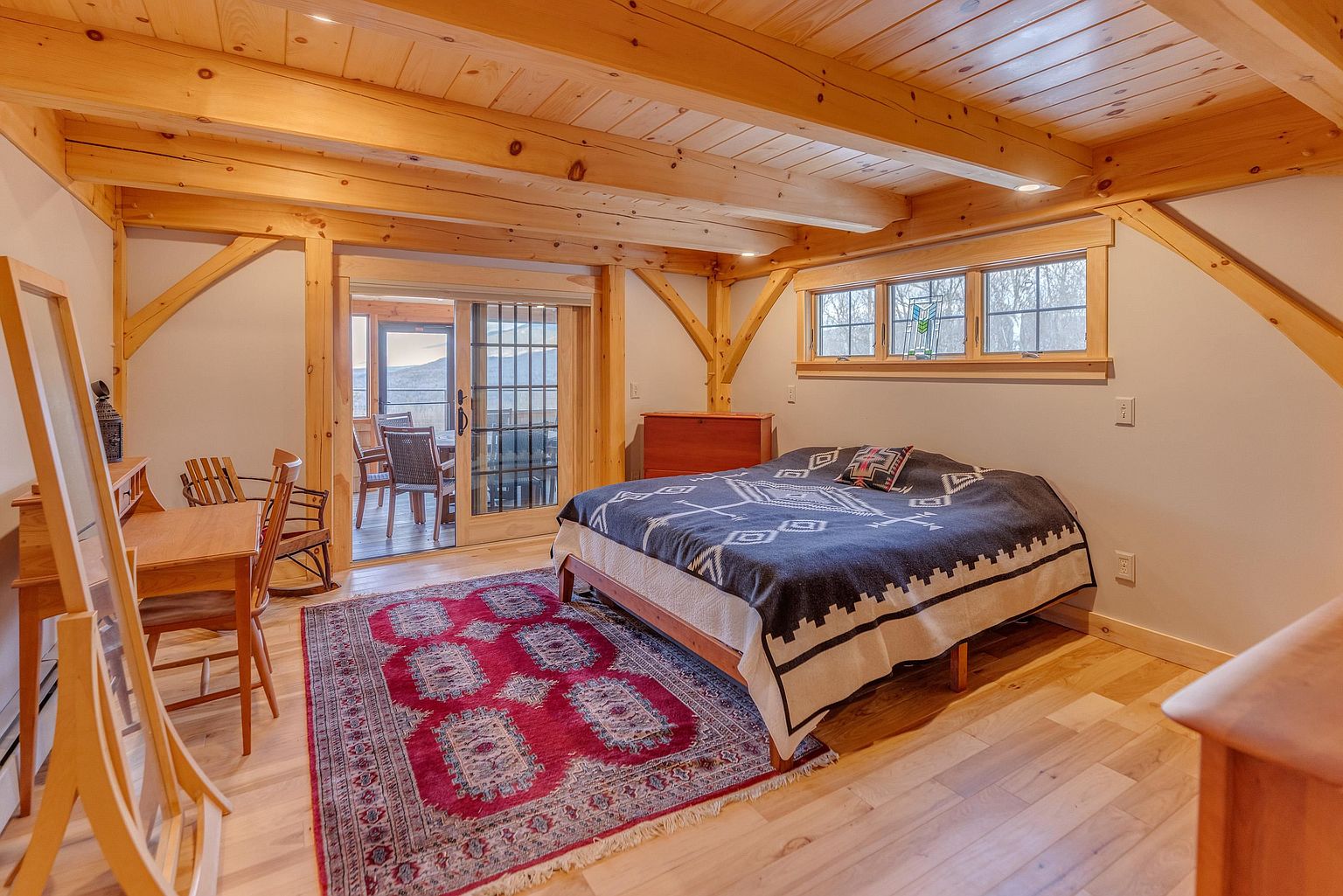 This rustic bedroom features exposed timber beams and light wood flooring, creating a warm and inviting atmosphere. A large bed with a patterned textile cover sits centrally, complemented by a vibrant, traditional-style area rug and a small wooden desk area. Large sliding glass doors provide a view into an adjacent sunroom, enhancing the sense of space and natural light within the room.
