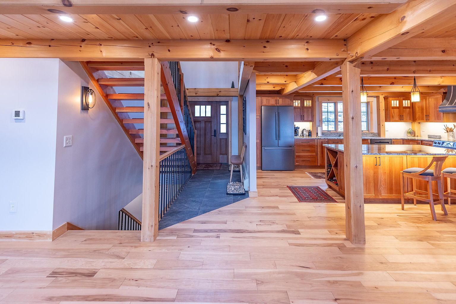 This open-concept interior features a warm, rustic aesthetic with exposed timber beams and light wood flooring throughout. The perspective captures a transition space that connects a slate-tiled entryway and staircase to a spacious kitchen area, highlighted by a large island and stainless steel appliances. The design emphasizes natural textures and an airy, inviting atmosphere suitable for a modern mountain home.