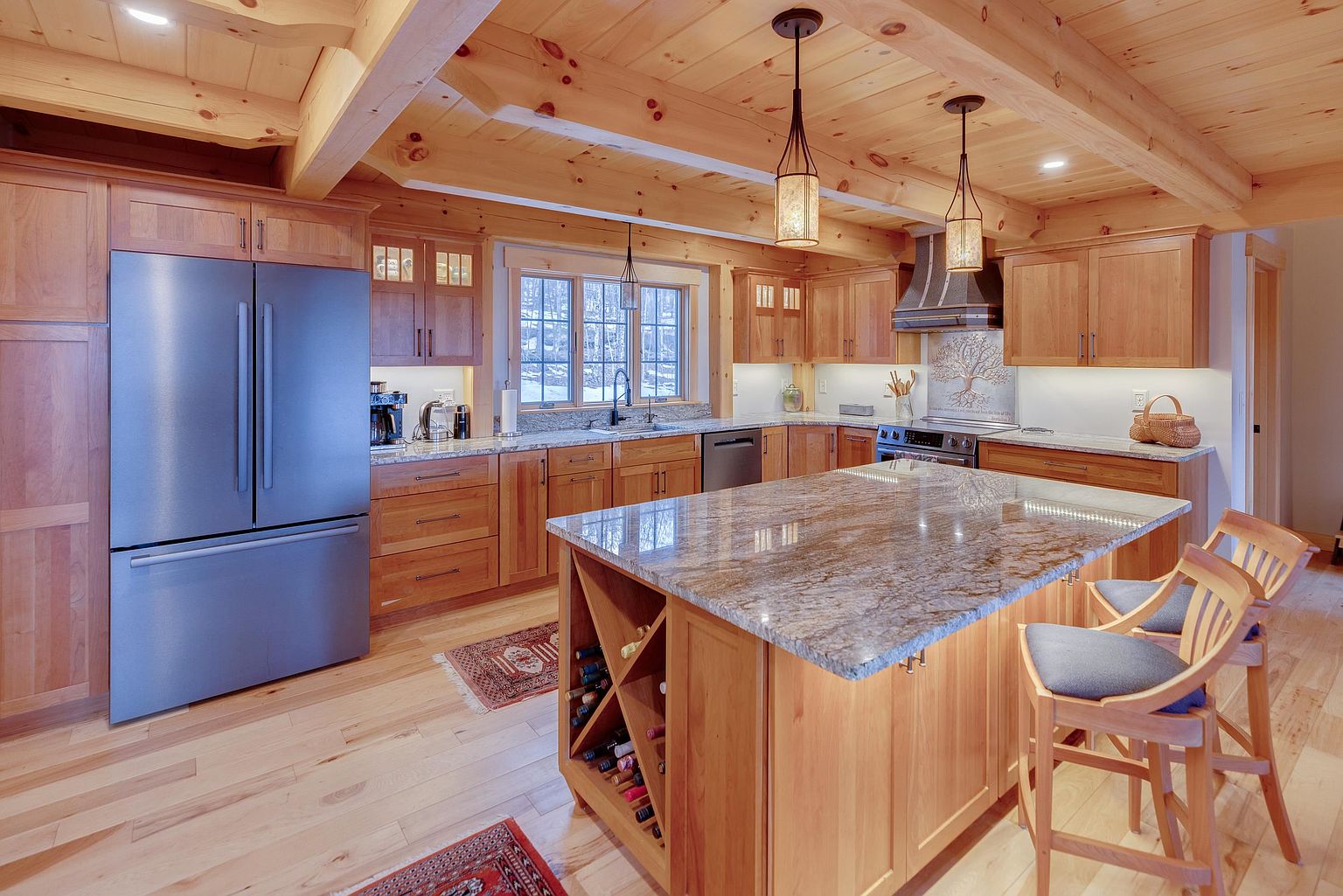 This warm and inviting kitchen features extensive light wood cabinetry, exposed ceiling beams, and a large central island topped with a polished stone surface. The space is equipped with stainless steel appliances, including a large refrigerator and a range with a decorative metal hood, while two pendant lights hang above the island. The room is accented by patterned rugs on the light wood flooring and a window over the sink that provides a view of the outdoors.
