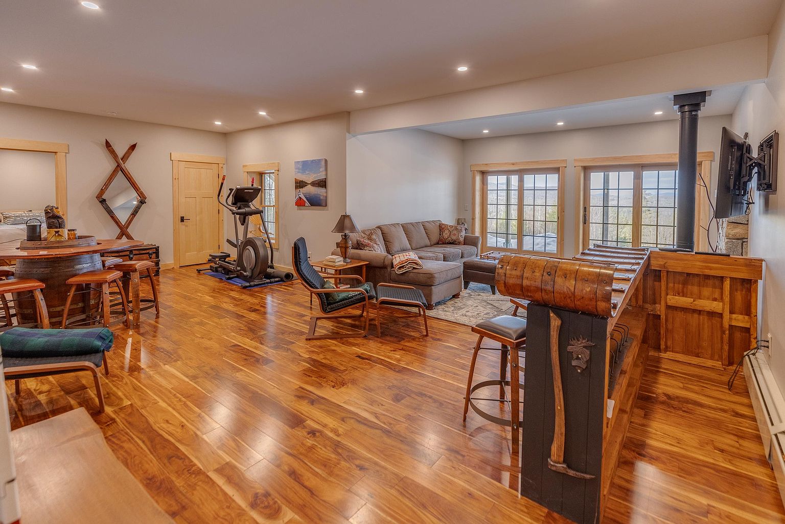 This spacious, open-concept basement features warm wood flooring and a versatile layout that combines a lounge area, a home gym, and a rustic bar. The living space is anchored by a comfortable sectional sofa and a leather armchair, while the bar area is uniquely crafted from a vintage-style sled. Large windows provide ample natural light and scenic views, creating an inviting atmosphere for relaxation and recreation.