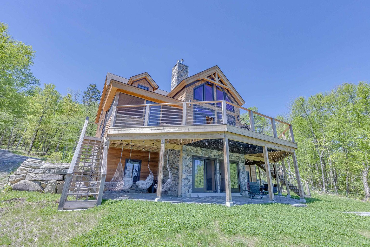 This rear view of a rustic-style home showcases a multi-level design with a large wooden deck overlooking a stone-paved patio. The lower level features a stone-clad exterior wall with sliding glass doors and hanging chairs, while the upper deck is enclosed by a modern cable railing. Surrounded by lush green trees and a natural landscape, the home presents a serene and inviting outdoor living space.