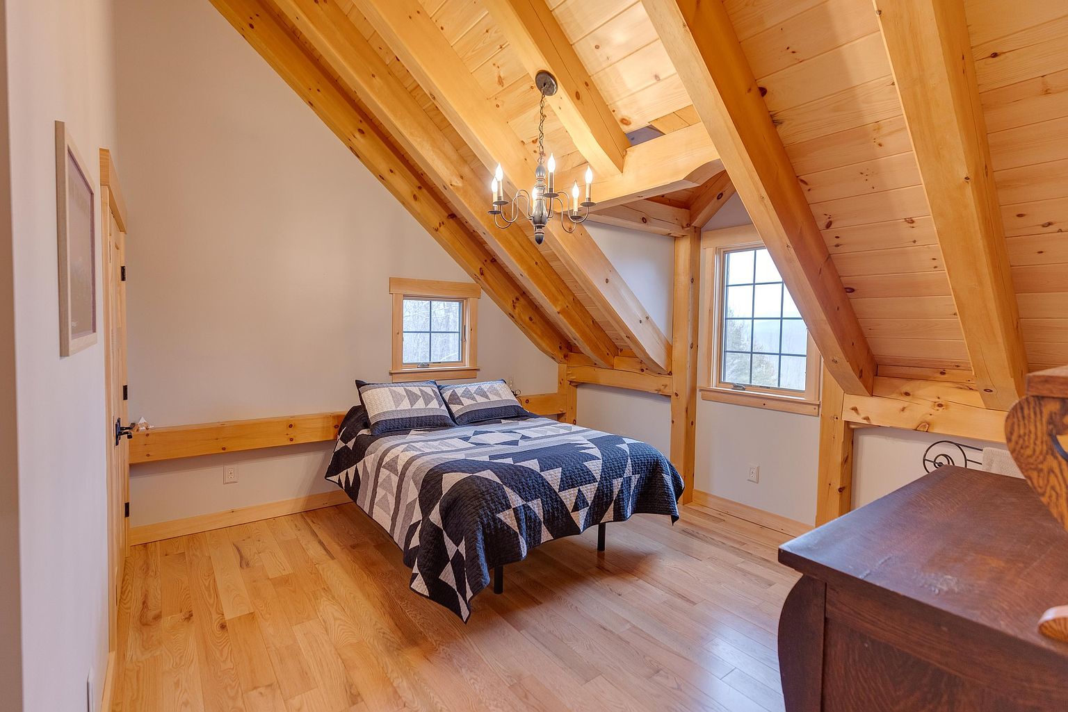 This cozy bedroom is located in an attic space, characterized by exposed timber beams and a vaulted ceiling finished with light wood planks. A bed with a geometric blue and white quilt sits centrally, flanked by two windows that provide natural light and views of the outdoors. The room features warm-toned hardwood flooring and a rustic chandelier, creating a charming and inviting atmosphere.