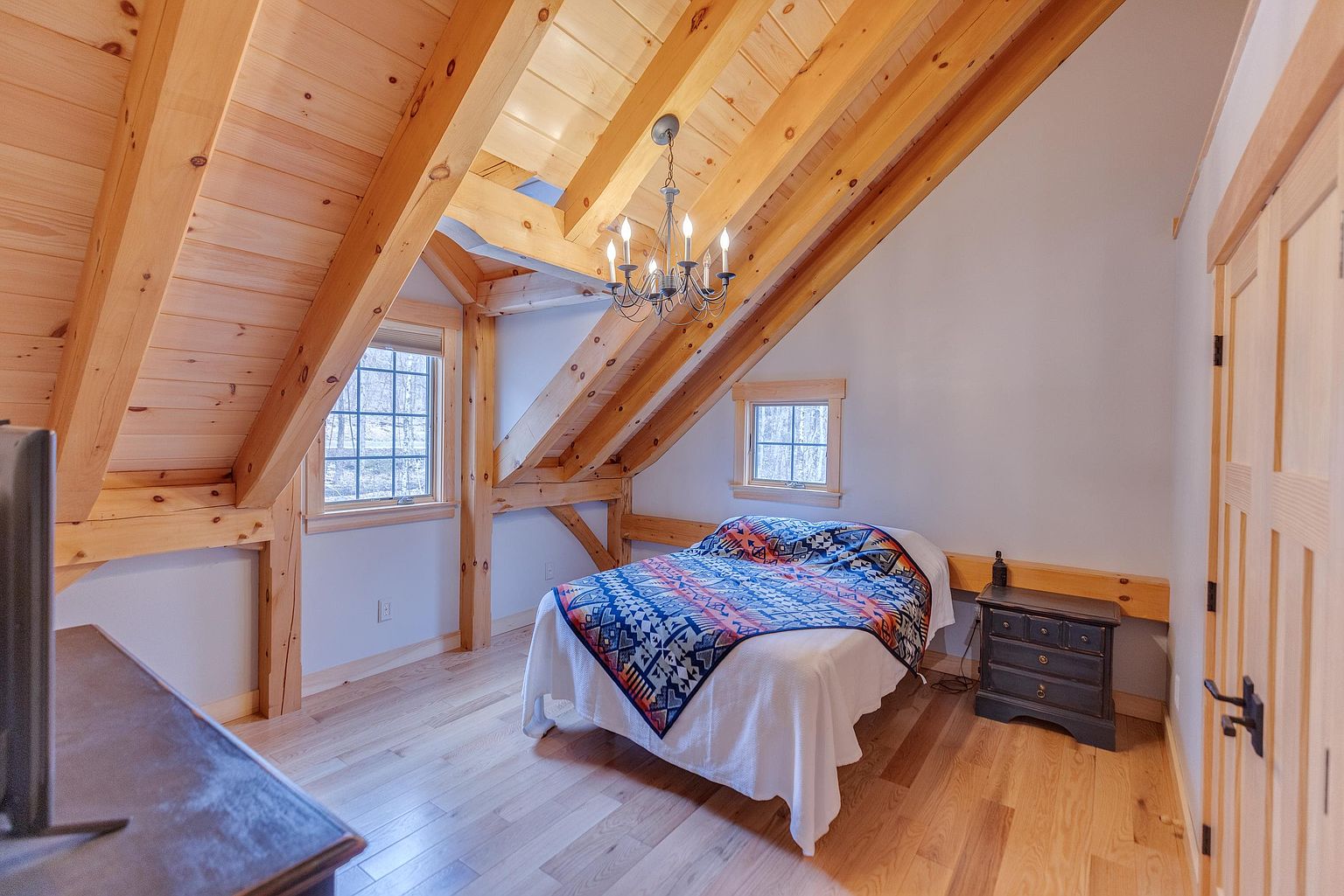This cozy bedroom features a rustic aesthetic with exposed timber beams and light wood flooring under a vaulted ceiling. A bed with a vibrant, patterned quilt serves as the focal point, complemented by a dark wood nightstand and a classic chandelier. The room is illuminated by natural light from two windows, creating a warm and inviting atmosphere.