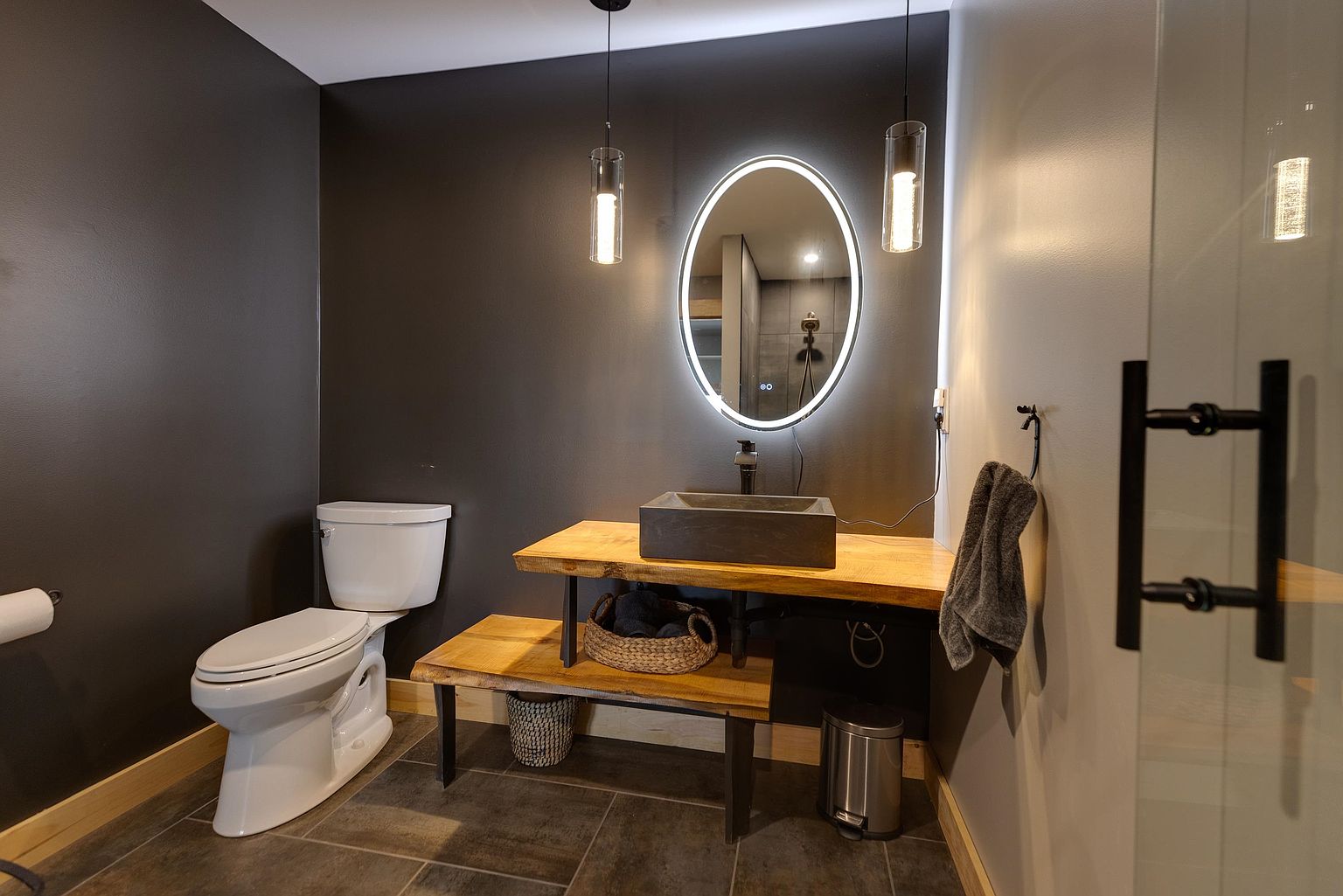 This modern bathroom features a striking live-edge wood vanity with a dark stone vessel sink and a backlit oval mirror. The space is anchored by dark walls and large-format floor tiles, creating a sophisticated, moody aesthetic. A white toilet and minimalist pendant lighting complete the contemporary design, offering a clean and stylish powder room experience.