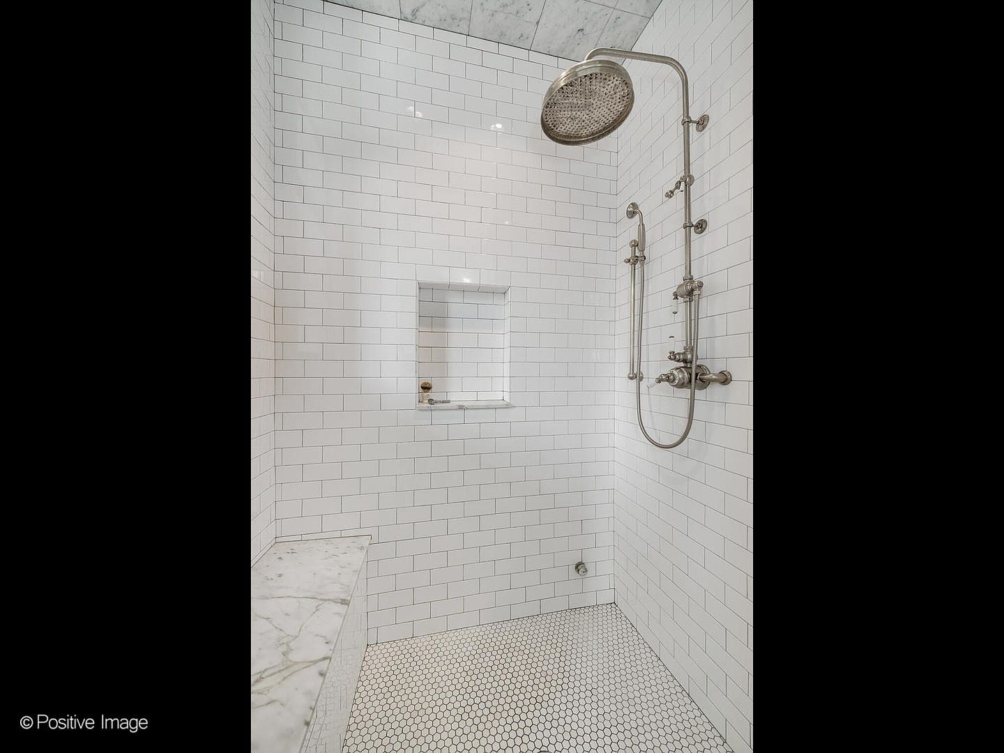 This walk-in shower features classic white subway tile walls and a contrasting hexagonal mosaic tile floor. A built-in stone bench sits to the left, while a polished metal shower system with a large rainfall head and a handheld sprayer is mounted on the right wall. A recessed niche provides convenient storage, and the space is finished with a stone-tiled ceiling, creating a clean and timeless aesthetic.