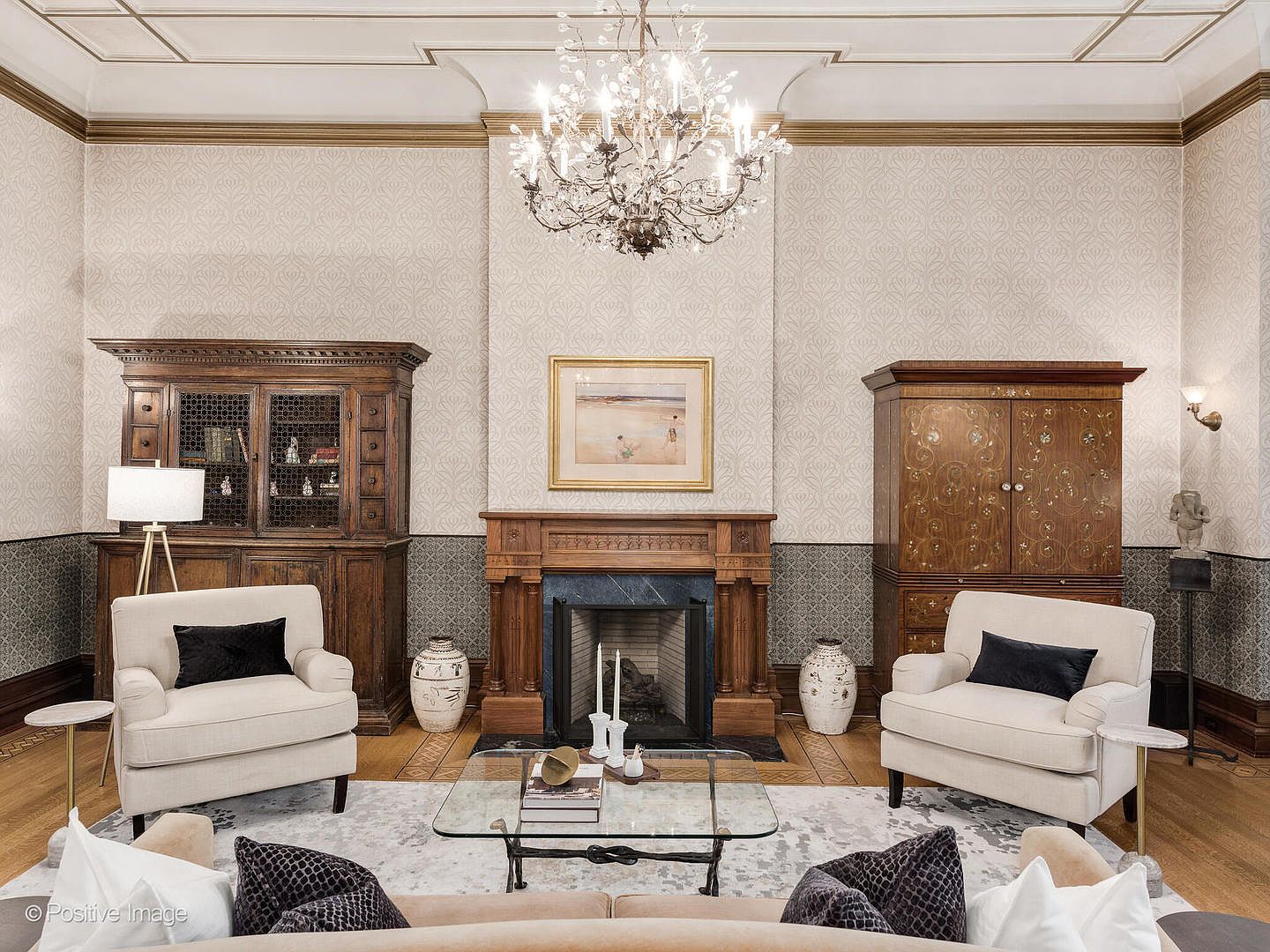 This elegant living room features a symmetrical layout centered around a traditional fireplace with a wood mantel and a dark stone surround. Two cream-colored armchairs with dark accent pillows flank the fireplace, while a glass-topped coffee table sits on a patterned area rug. The room is adorned with a crystal chandelier, ornate wallpaper, and two large wooden cabinets, creating a sophisticated and formal atmosphere.
