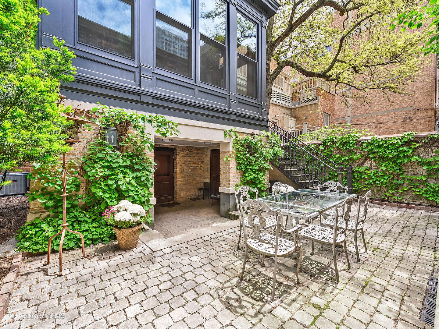 This charming, private patio features a classic cobblestone floor and is framed by lush climbing ivy against a brick wall. A glass-topped dining set with ornate metal chairs sits in the center, while a recessed entryway leads into the home beneath a dark-framed, multi-pane window structure. A metal staircase ascends along the side, adding architectural interest to this serene, secluded outdoor living space.