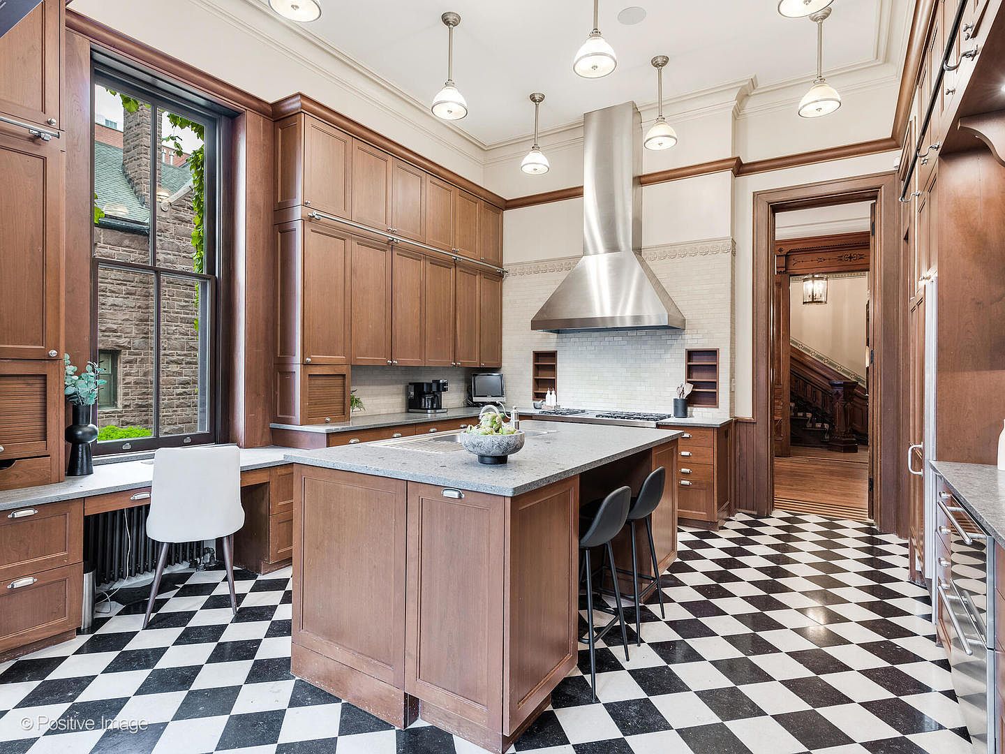 This elegant kitchen features warm-toned wood cabinetry, a large central island with stone countertops, and a striking black-and-white checkered floor. A stainless steel range hood serves as a focal point above the cooktop, while multiple pendant lights provide bright illumination throughout the space. The room includes a built-in desk area by a large window and offers a clear view into an adjacent hallway with a grand staircase.