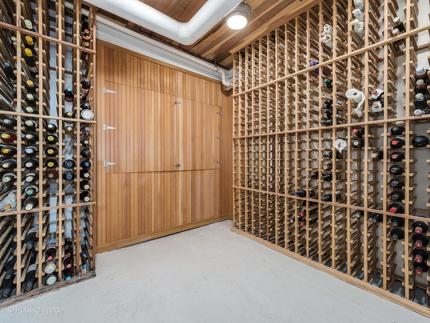 This dedicated wine cellar features extensive custom wood racking systems that span the walls, providing ample storage for a large collection. A central wall is finished with vertical wood paneling, adding a warm, textured element to the space. The room is illuminated by a single ceiling-mounted light fixture and has a simple, neutral floor, creating a functional and organized environment for wine storage.