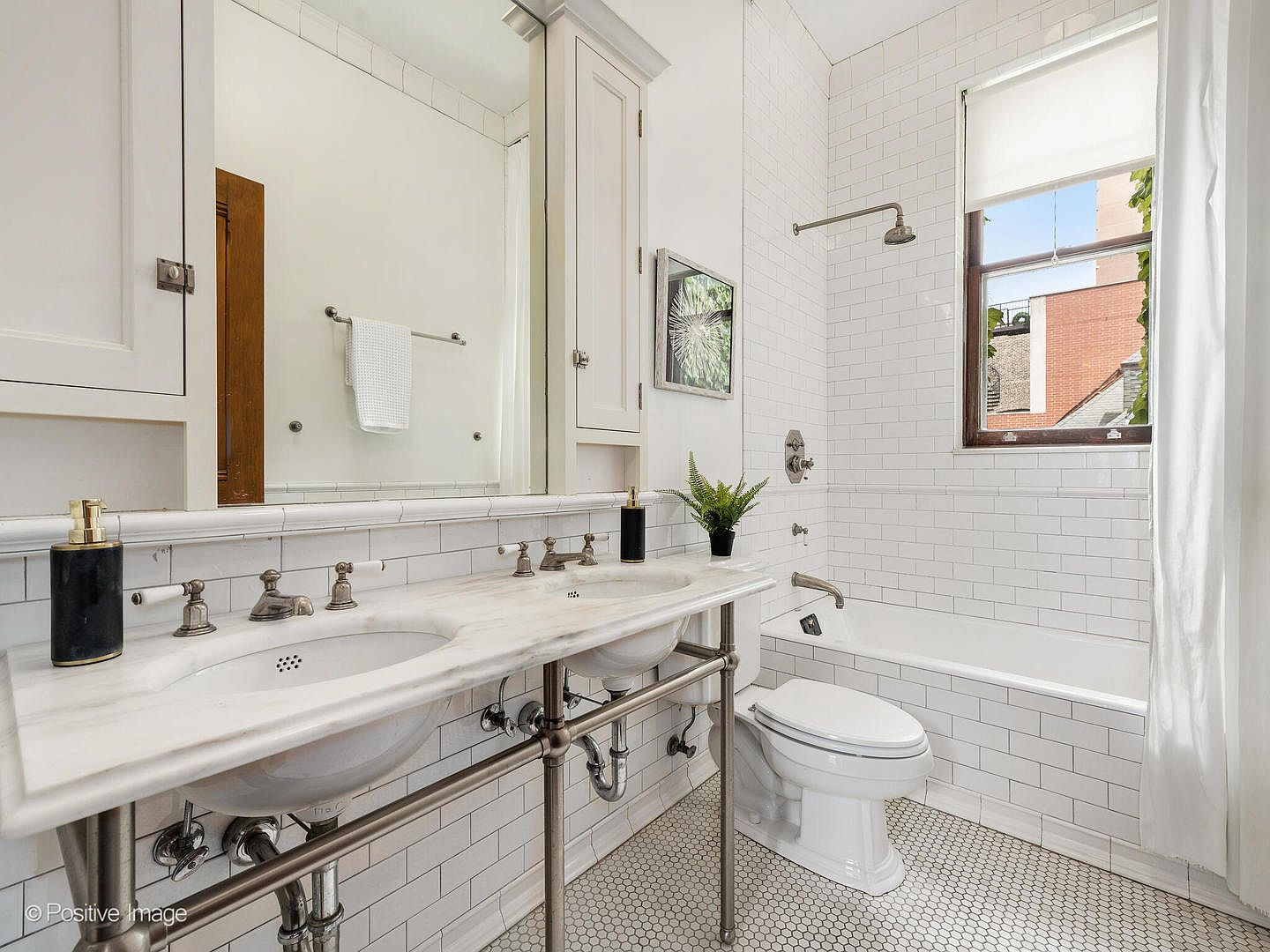 This bright, classic bathroom features a double-sink vanity with a stone countertop and exposed metal plumbing fixtures. The space is finished with white subway tile on the walls and a small-scale hexagonal tile on the floor, creating a clean, timeless aesthetic. A combination shower and bathtub is positioned under a window, and a large mirror above the vanity enhances the sense of light and space.