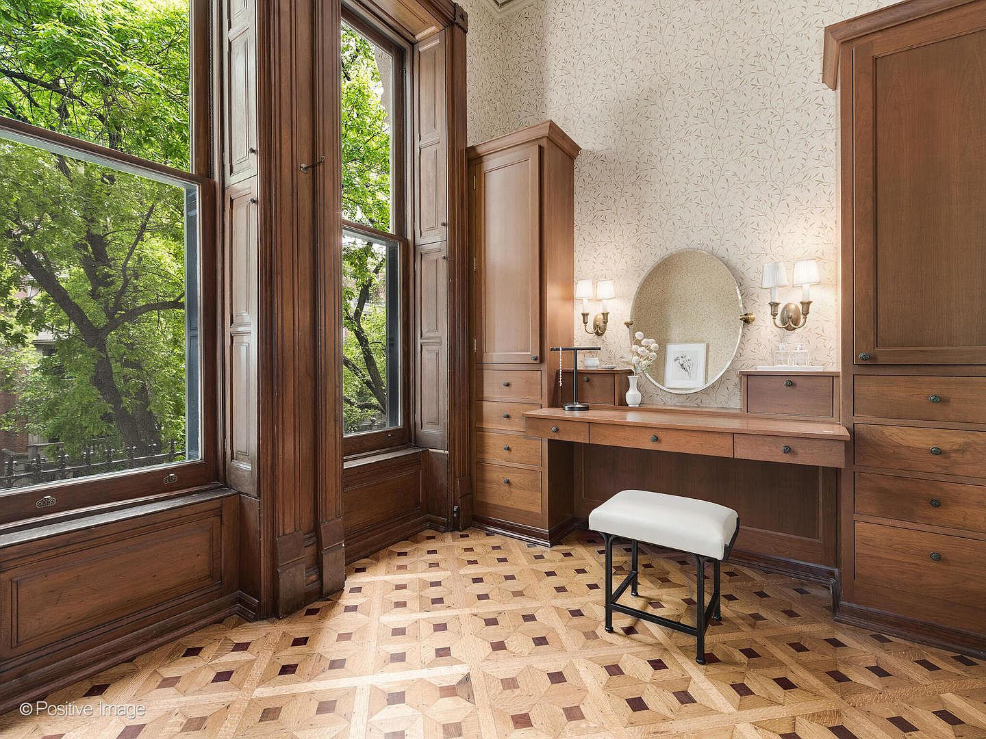 This elegant dressing area features custom built-in wood cabinetry surrounding a central vanity with a round mirror and wall-mounted sconces. The room is highlighted by intricate parquet flooring and tall windows framed in rich wood trim that provide a view of lush greenery outside. A simple white stool sits at the vanity, creating a refined and functional space within the home.