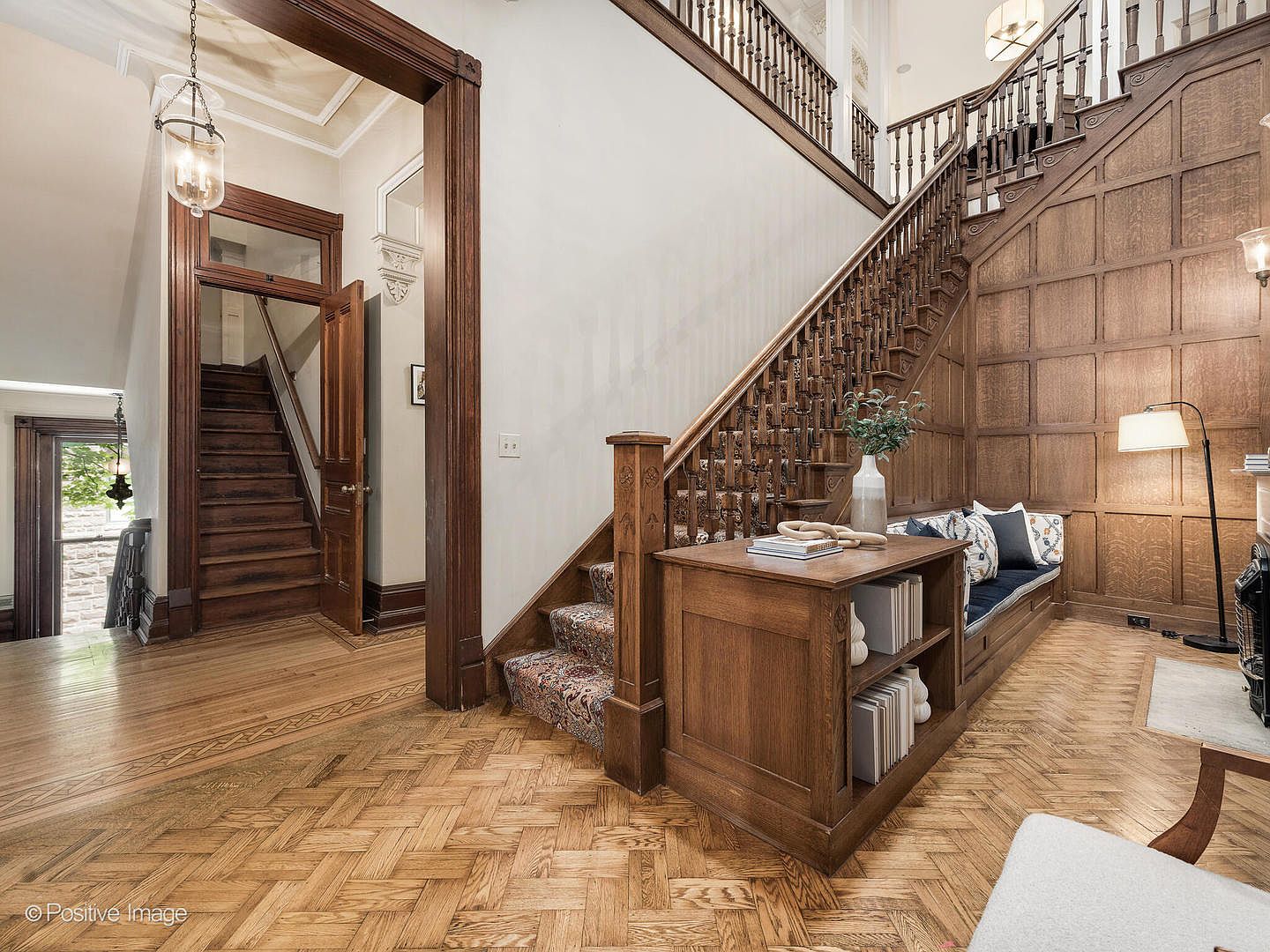 This grand entryway features a striking wooden staircase with intricate balusters and a built-in bench seat nestled against wood-paneled walls. The space showcases rich wood tones, a herringbone-patterned wood floor, and a secondary staircase visible through a doorway in the background. The perspective is from the foyer, highlighting the architectural craftsmanship and the warm, inviting atmosphere of the home.