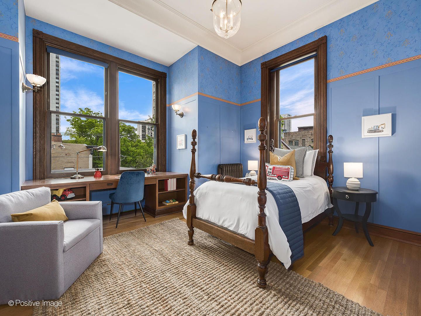 This charming bedroom features a classic four-poster wooden bed as the centerpiece, set against vibrant blue walls with decorative wallpaper accents. A built-in desk area spans the space beneath two large windows, providing a functional workspace, while a cozy armchair and a textured area rug add comfort to the room. The space is well-lit by a central pendant fixture and wall-mounted sconces, creating a warm and inviting atmosphere.
