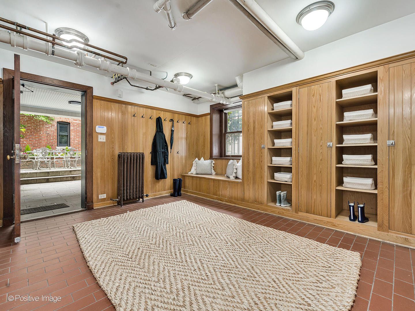 This mudroom or basement entryway features warm wood-paneled walls, built-in storage lockers with open shelving, and a long bench with decorative pillows. A large, textured area rug sits on the tiled floor, while an open doorway provides a view out to a brick-paved patio area. The space is illuminated by overhead lighting and includes exposed industrial-style piping along the ceiling, giving it a functional and rustic character.