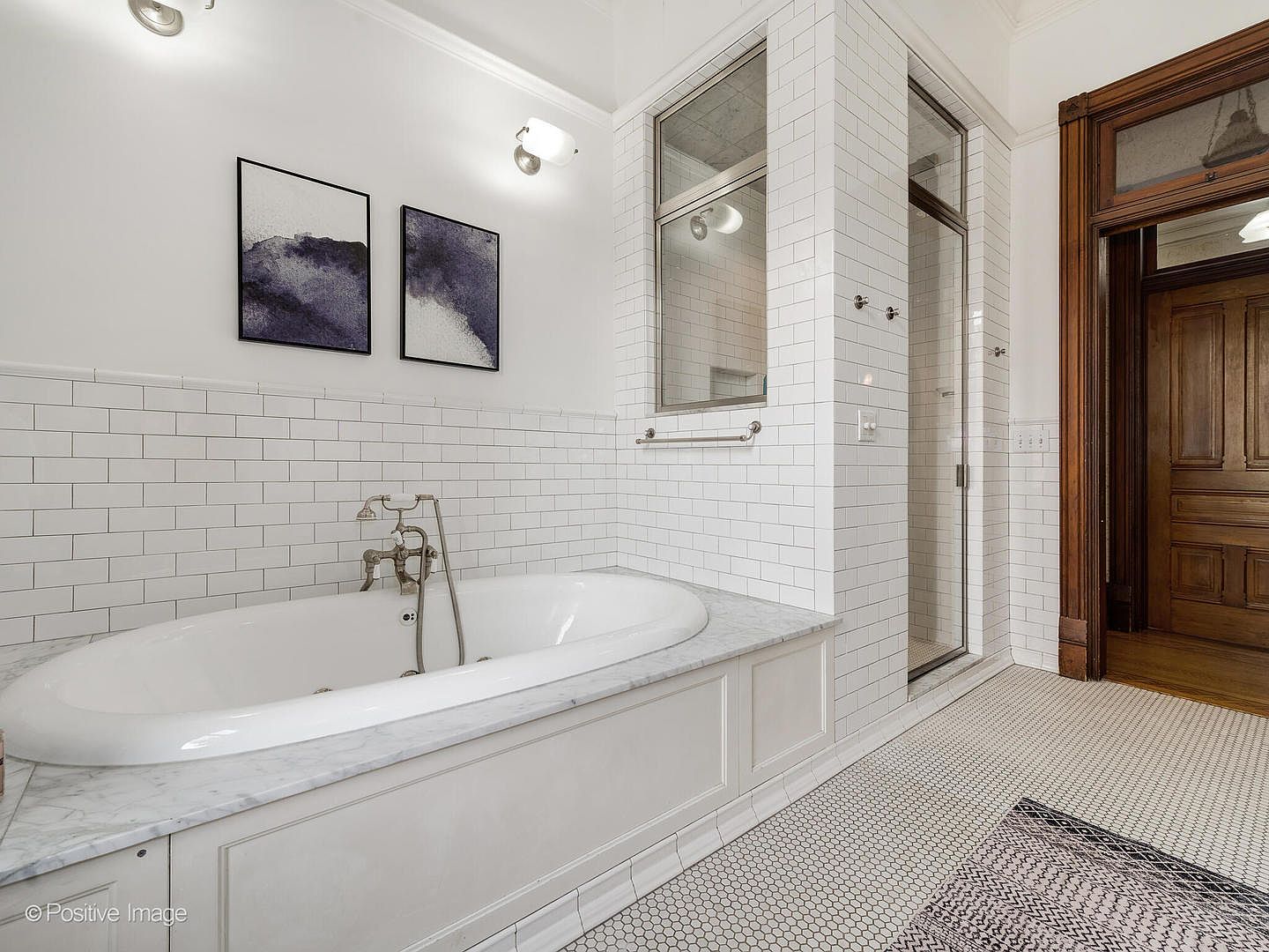 This elegant bathroom features a deep soaking tub set into a stone-topped vanity with white cabinetry. The walls are adorned with classic white subway tile, complemented by a glass-enclosed shower area and a patterned tile floor. A wooden door frame adds a touch of traditional warmth to the bright, clean aesthetic of the space.